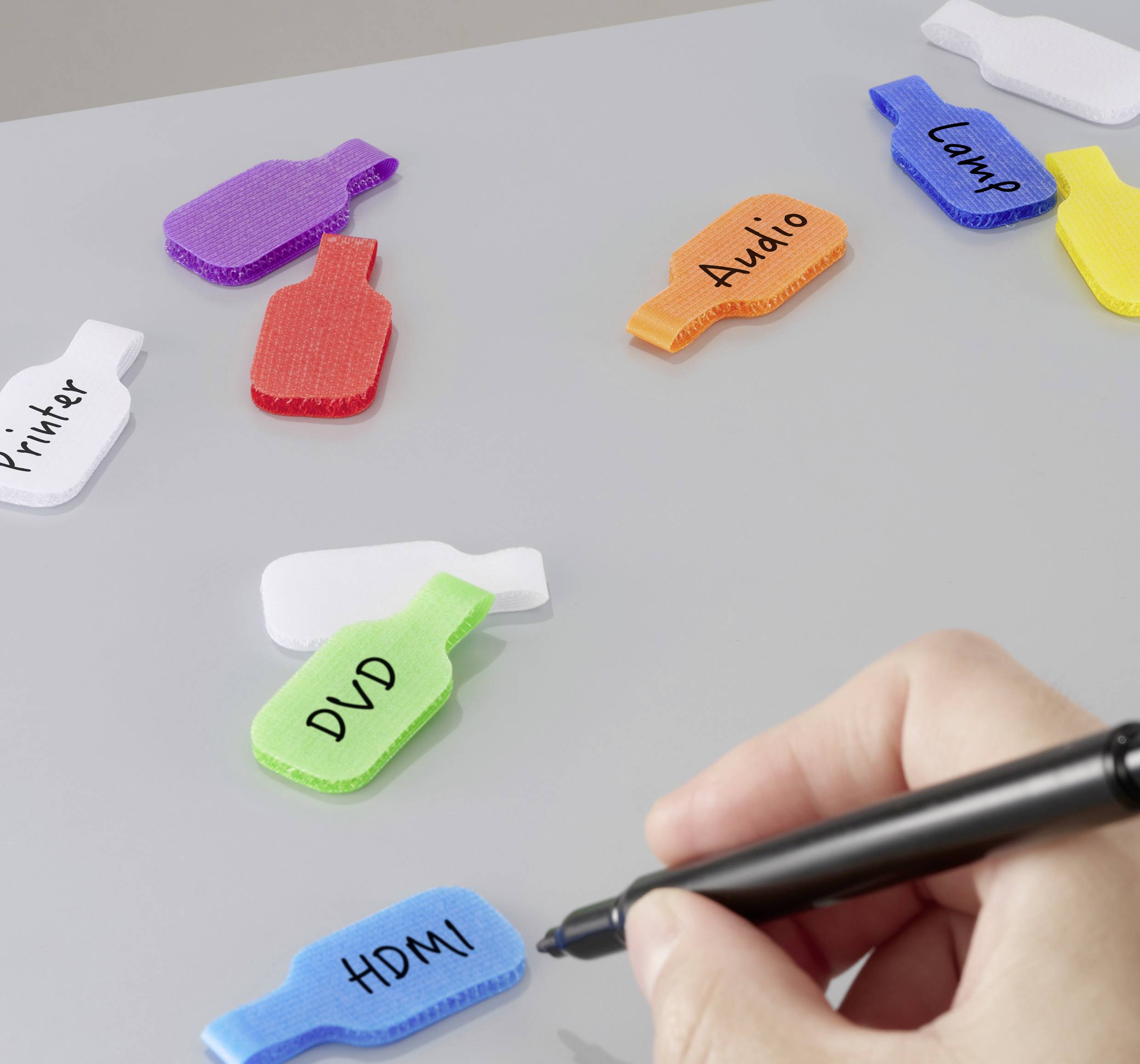 En hånd skriver på en lille hvid etiket med en kuglepen. I nærheden er farverige etiketter markeret på kabler: 'Printer,' 'DVD,' 'HDMI,' 'Lyd,' 'Lejr.'