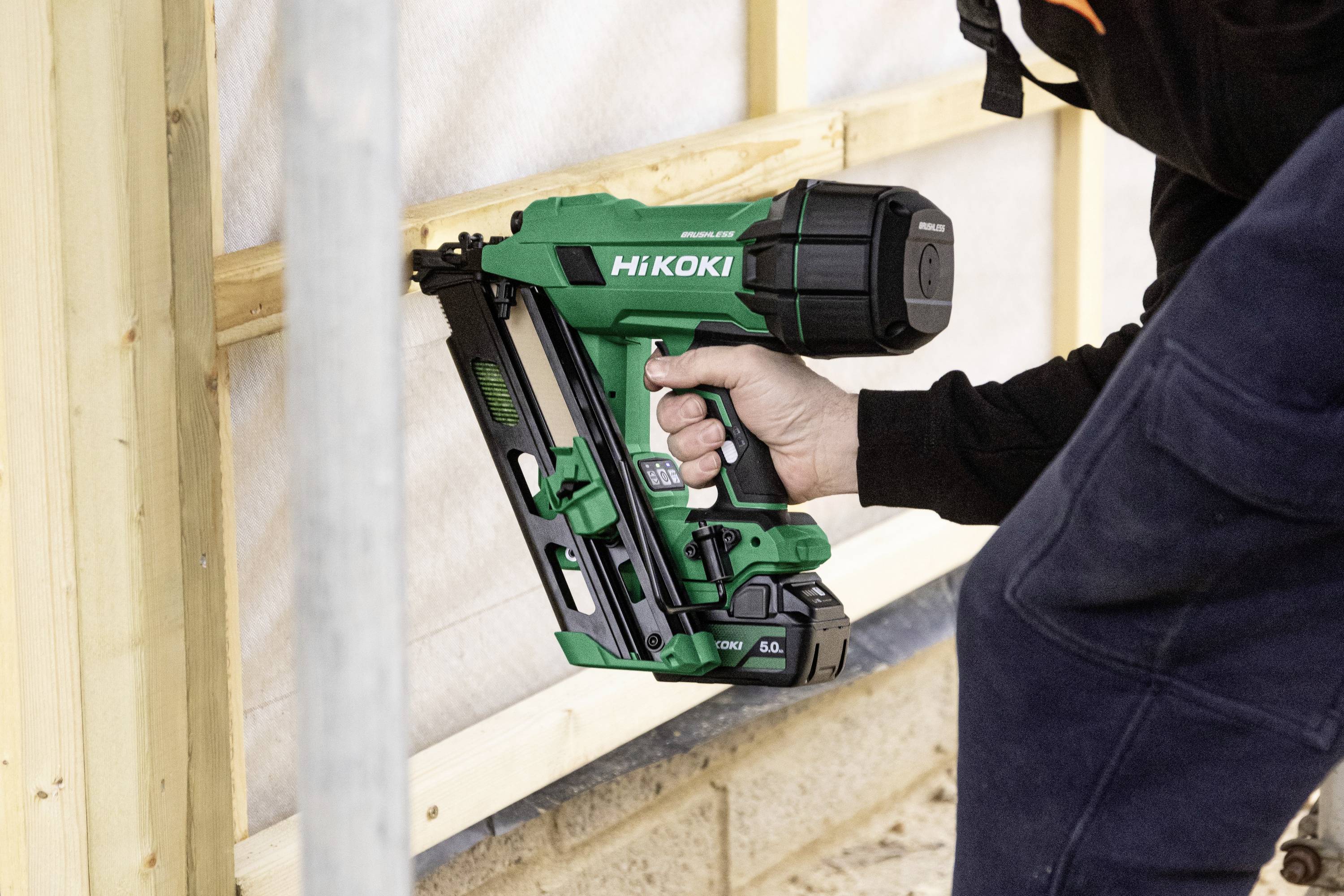 En person bruger en grøn HiKOKI sømpistol til at fastgøre træramme på en udendørs byggeplads.