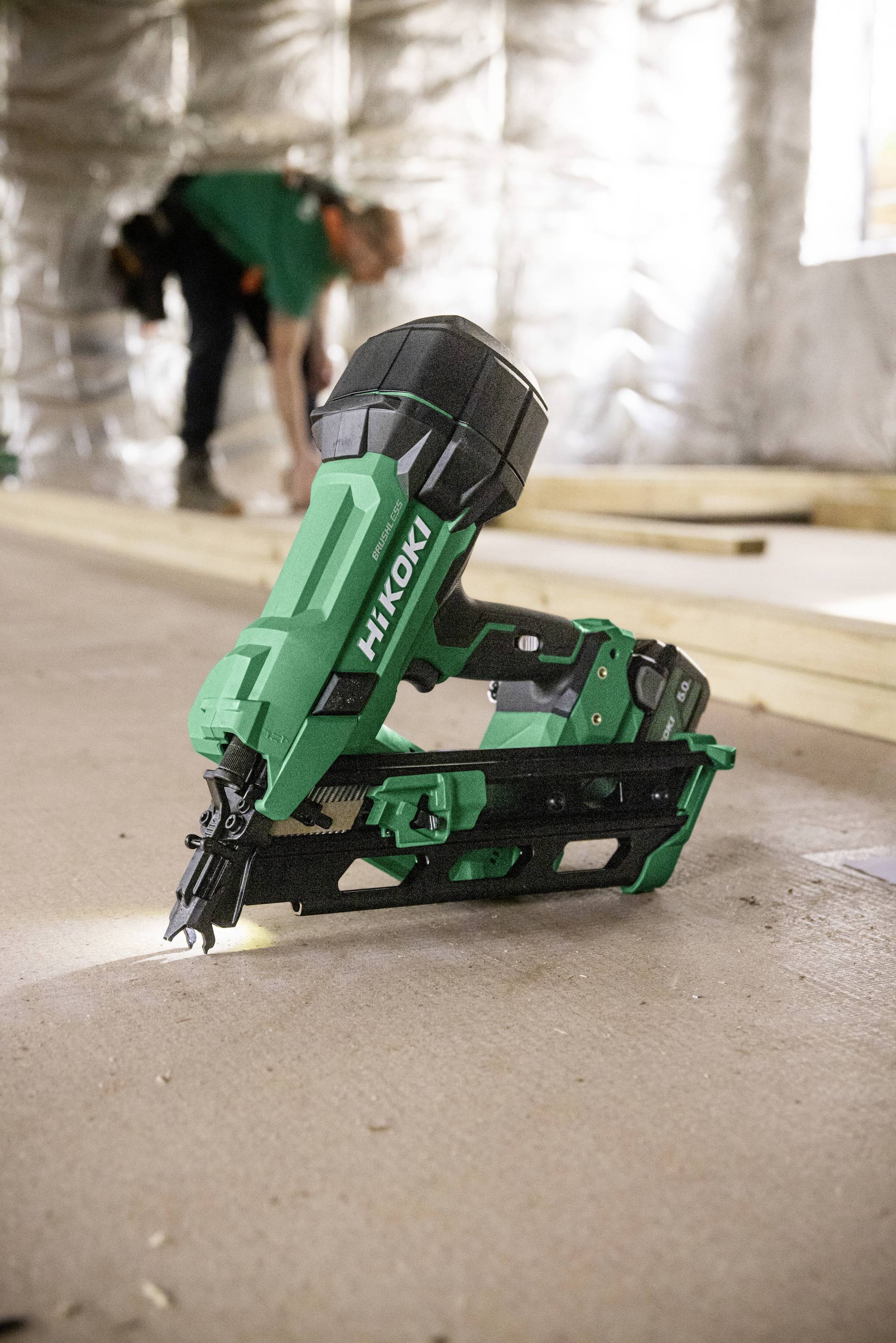En grøn HiKOKI trådløs sømpistol er placeret med bygningsarbejdere og træbjælker synlige i den slørede baggrund af en byggeplads.