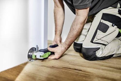 En person bruger et elektrisk værktøj til at skære i bunden af en dør, mens vedkommende sidder på knæ på et trægulv, med fokus på hjemmeforbedring eller renovering.