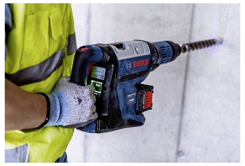 En person iført en gul sikkerhedsvest og handsker bruger en Bosch-boremaskine på en betonvæg.