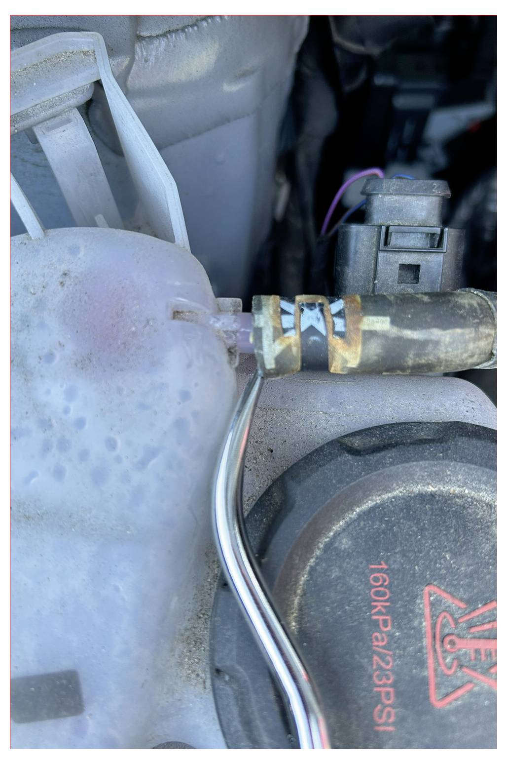 Et nærbillede af en bilmotorkomponent, der viser en rusten metalspænding på en slange forbundet til en hvid plastikbeholder. Et låg er mærket '160KPa/23PSI'.