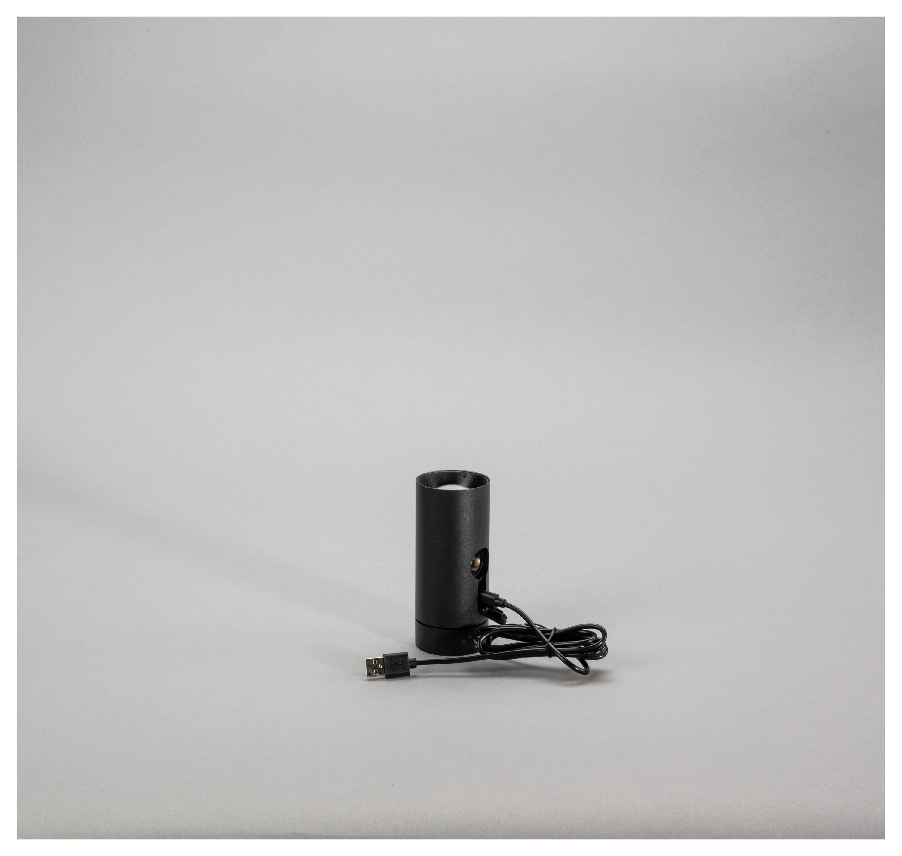 'En sort cylinderformet enhed med et USB-kabel tilsluttet, placeret på en ensfarvet grå overflade.'
