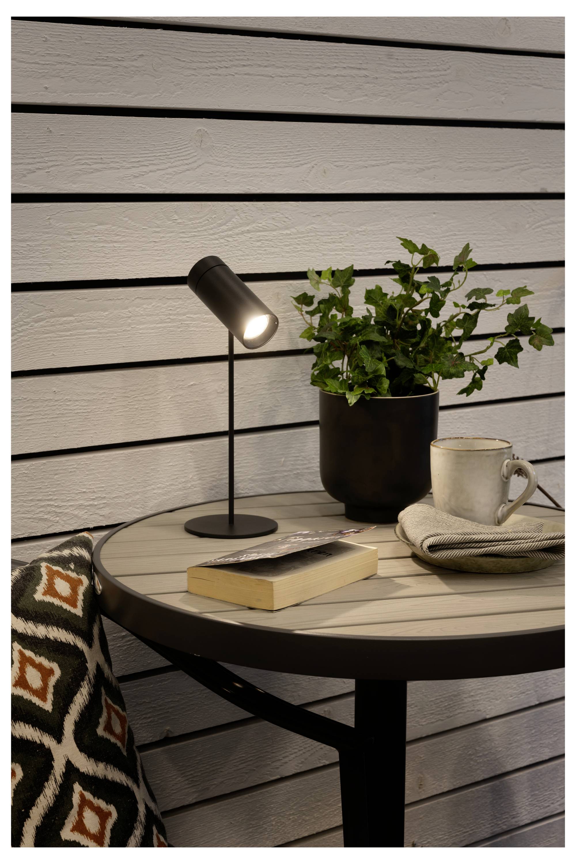 Et lille rundt bord med en lampe, plante, bog og krus ved siden af en stol. Hyggelig læseniche opsat foran en panel-beklædt væg.