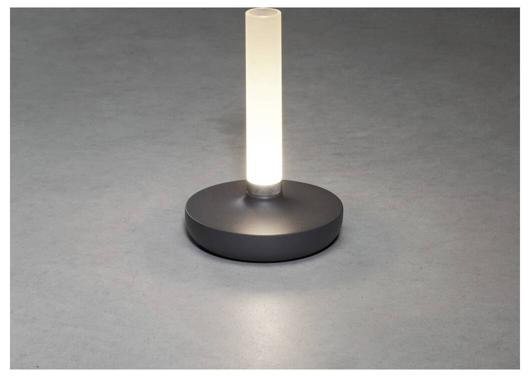 Et lille, moderne bordlampe med en cylinderformet lyskilde og rund base udsender et blødt skær på en grå overflade.
