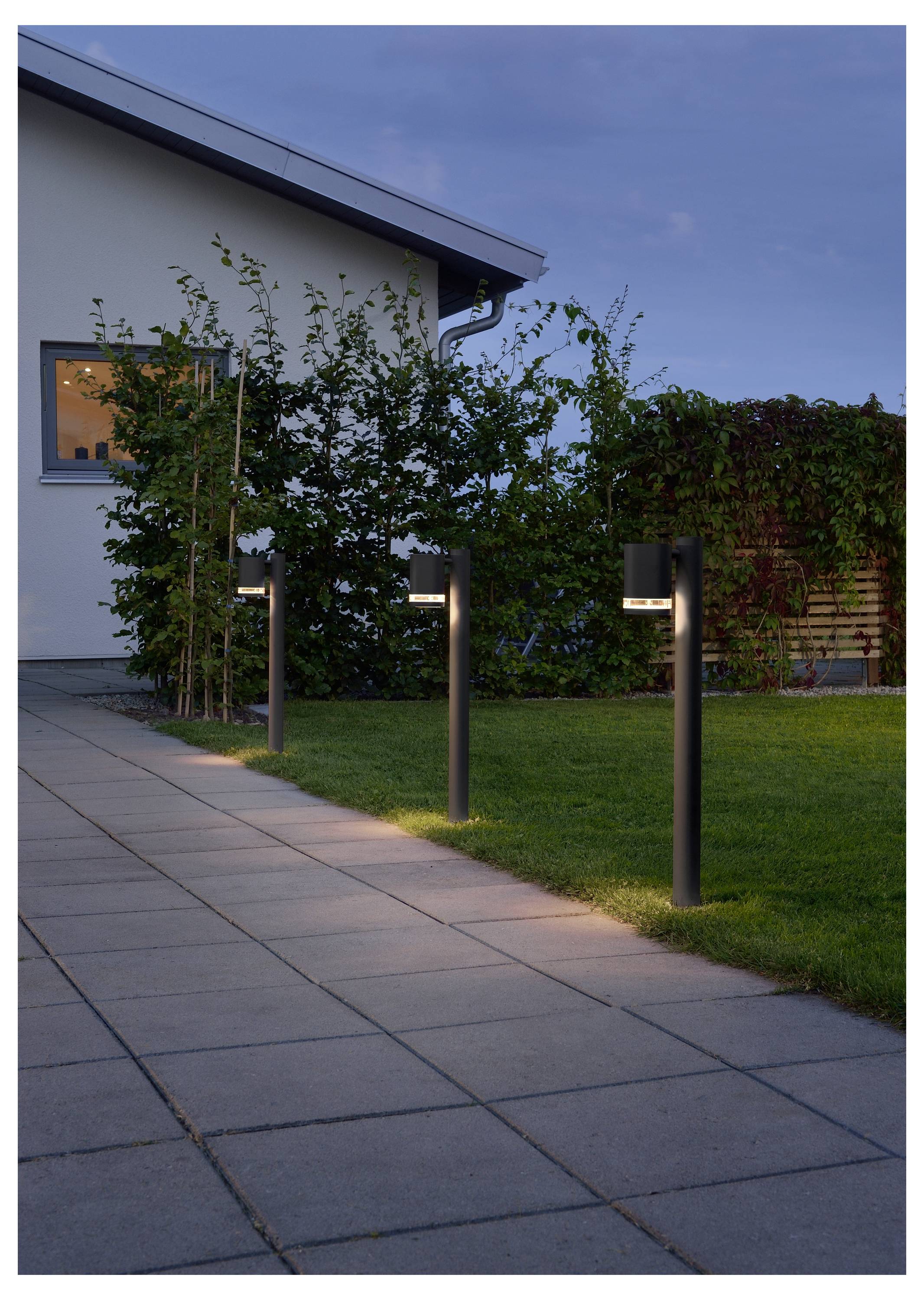 Udendørs stiplysene tændt ved skumringen ved siden af et moderne hus, der kaster et varmt lys på en flisebelagt sti omkranset af grønne planter.