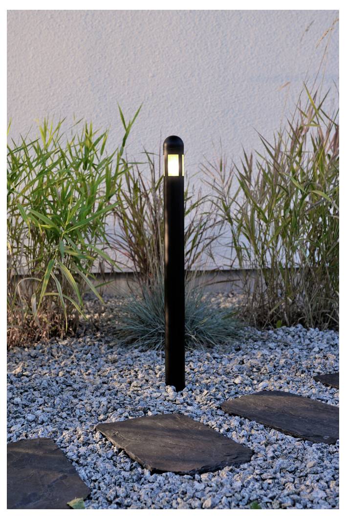 En sort udendørs pullertlampe oplyser en grusvej ved siden af trædesten og planter op mod en lys væg.
