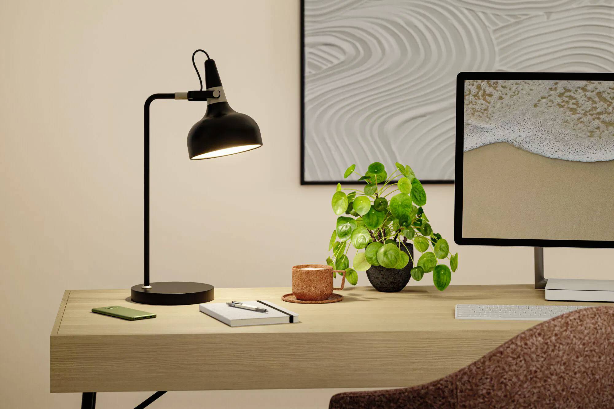 Et moderne skrivebord sat op med en sort lampe, en potteplante, en notesbog og en smartphone. En computer viser et strand-billede. Kunst pryder væggen.