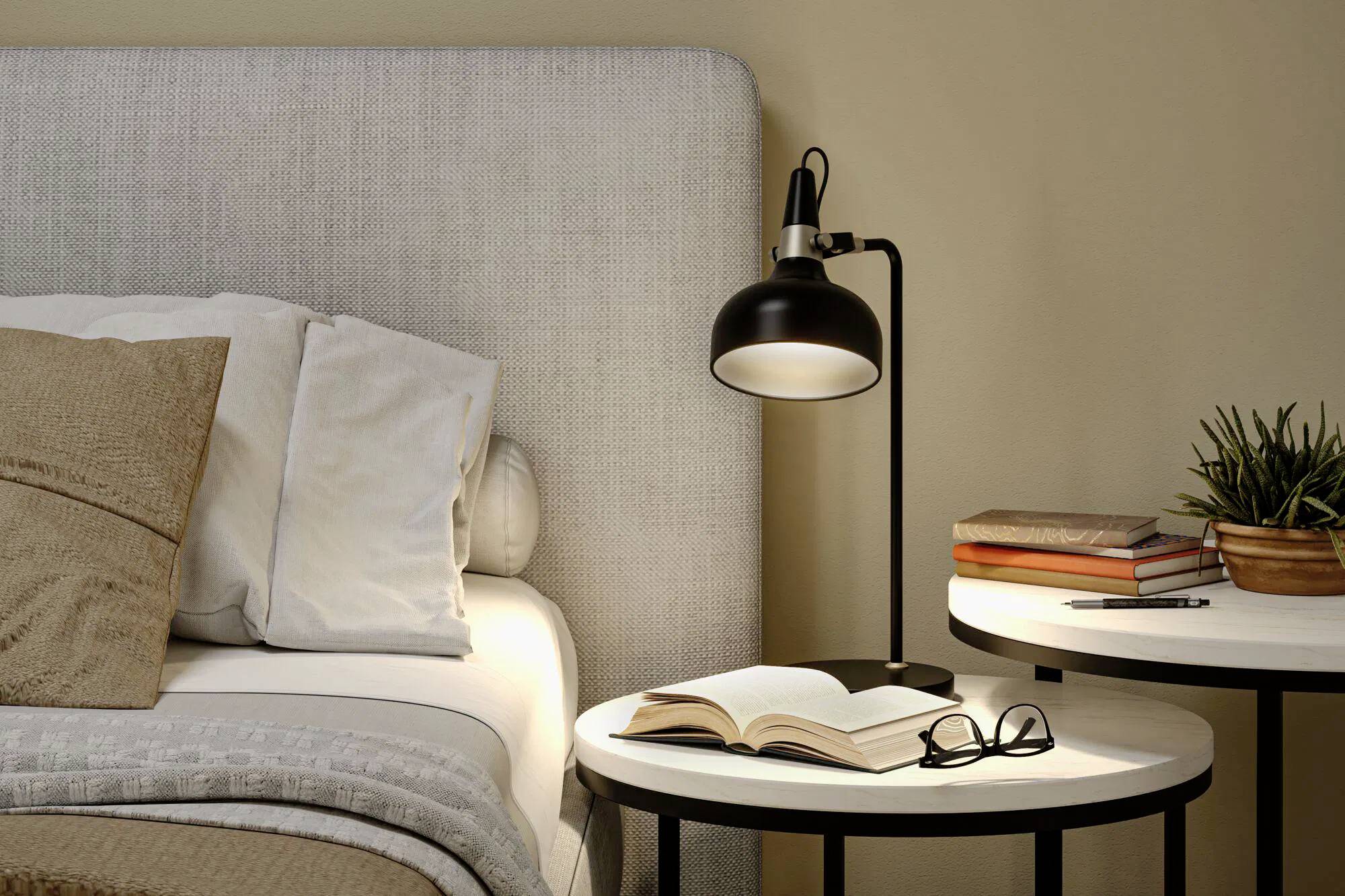 Et hyggeligt soveværelse har en seng med beige puder, et sengebord med en åben bog, briller og en moderne lampe, hvilket skaber en afslappet atmosfære.
