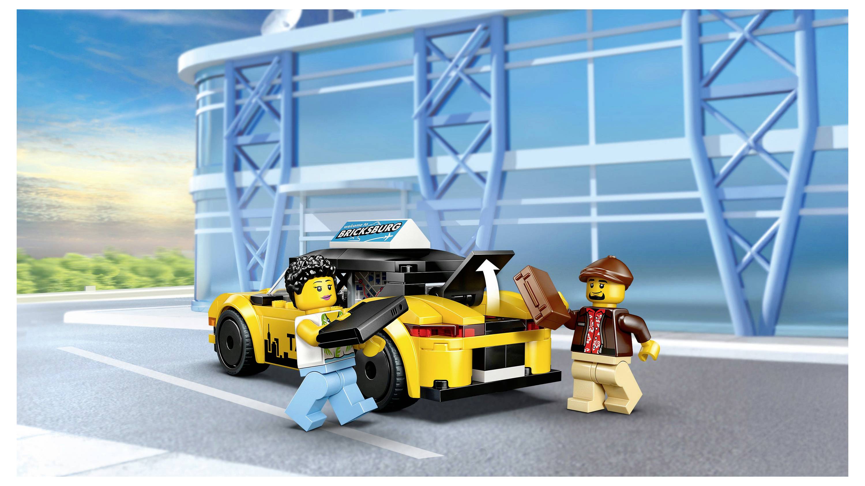 To legetøjsfigurer med kameraer står ved siden af en gul legetøjsbil uden for en bygning. Den blå himmel og moderne arkitektur danner baggrund.