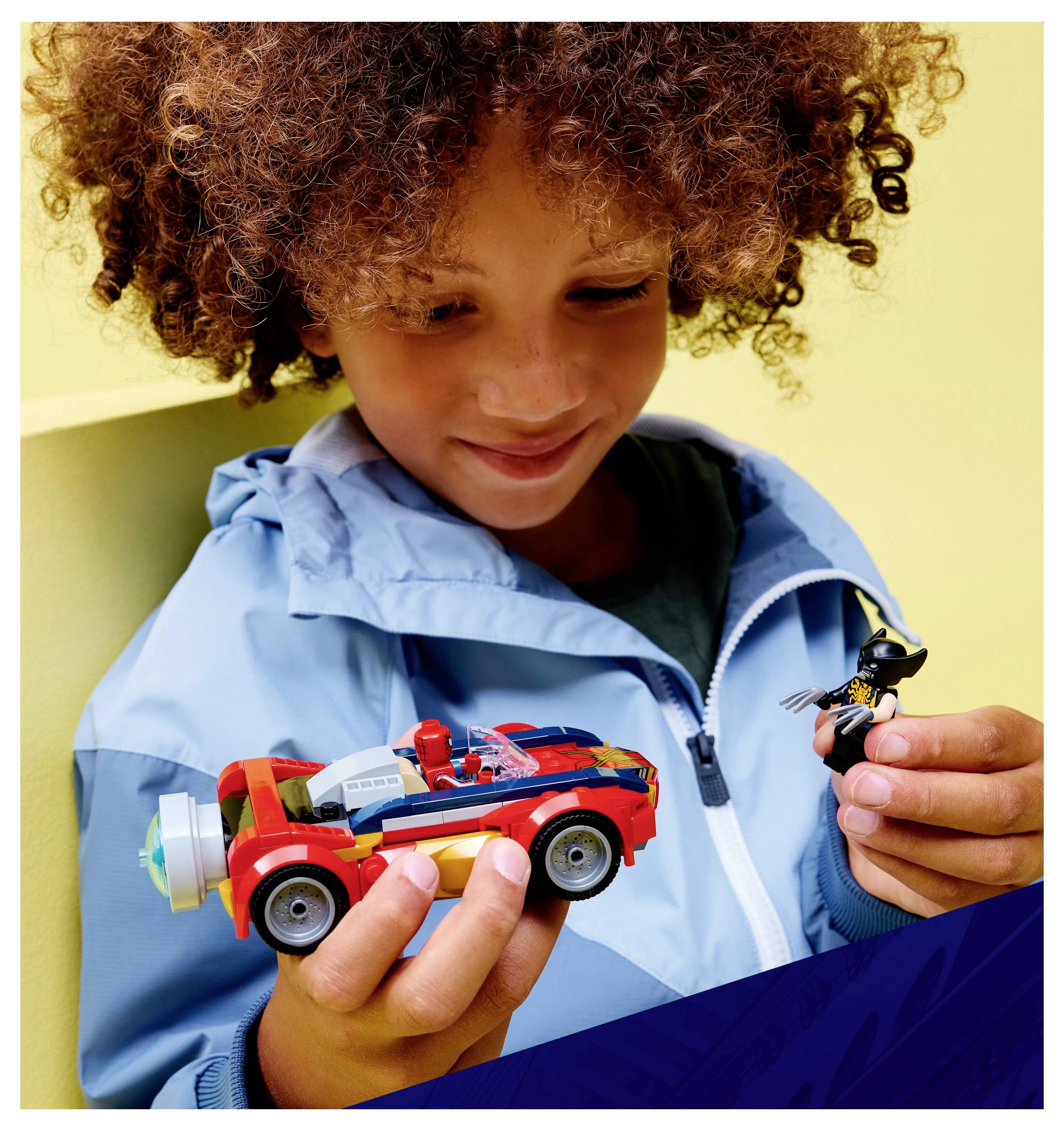 Et barn med krøllet hår, iført en blå jakke, smiler mens det holder en rød legebil i den ene hånd og en actionfigur i den anden, mod en gul baggrund.