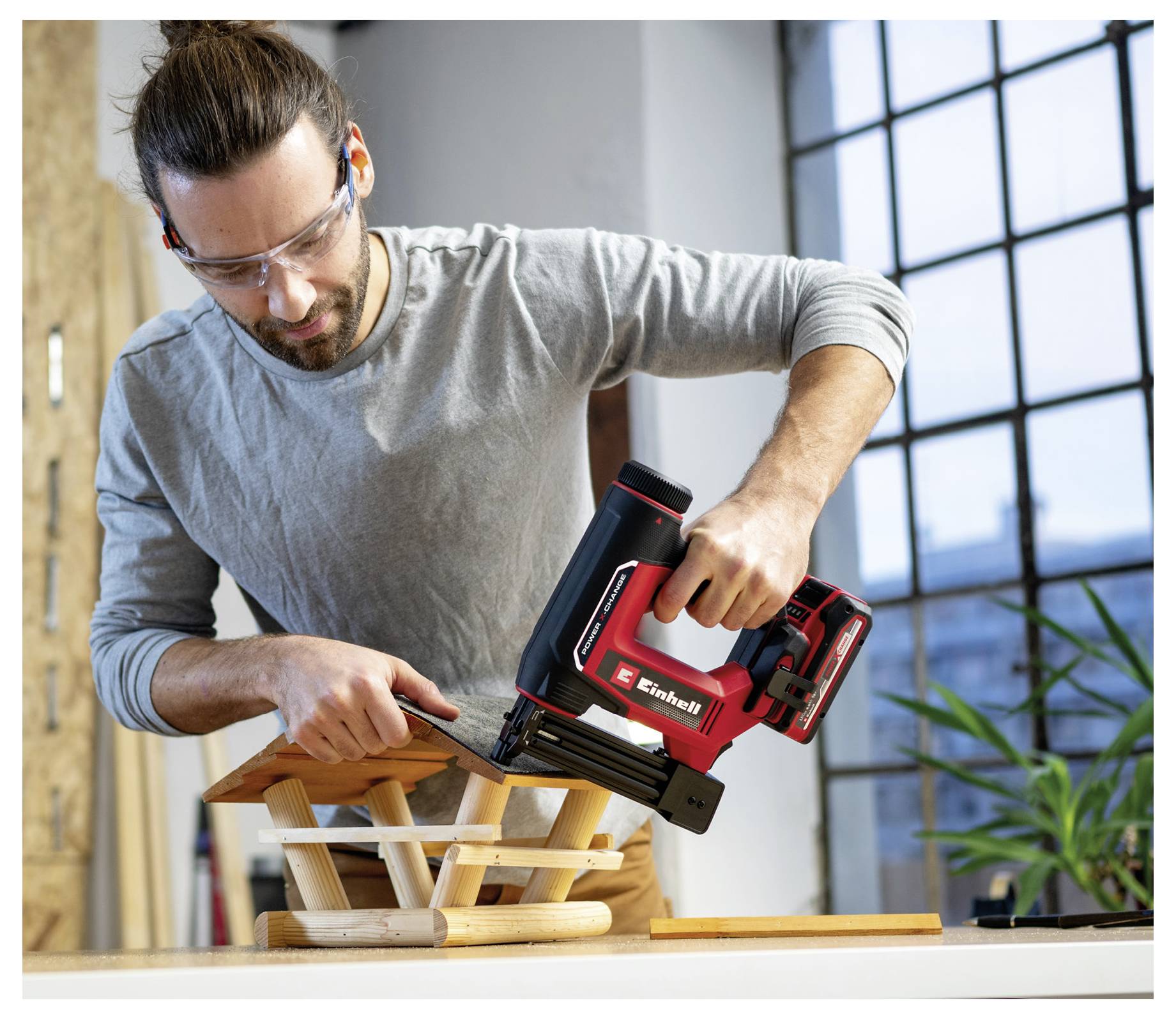 En person, der bærer sikkerhedsbriller, bruger et elværktøj til at samle en træstruktur indendørs, med et vindue og en plante i baggrunden.