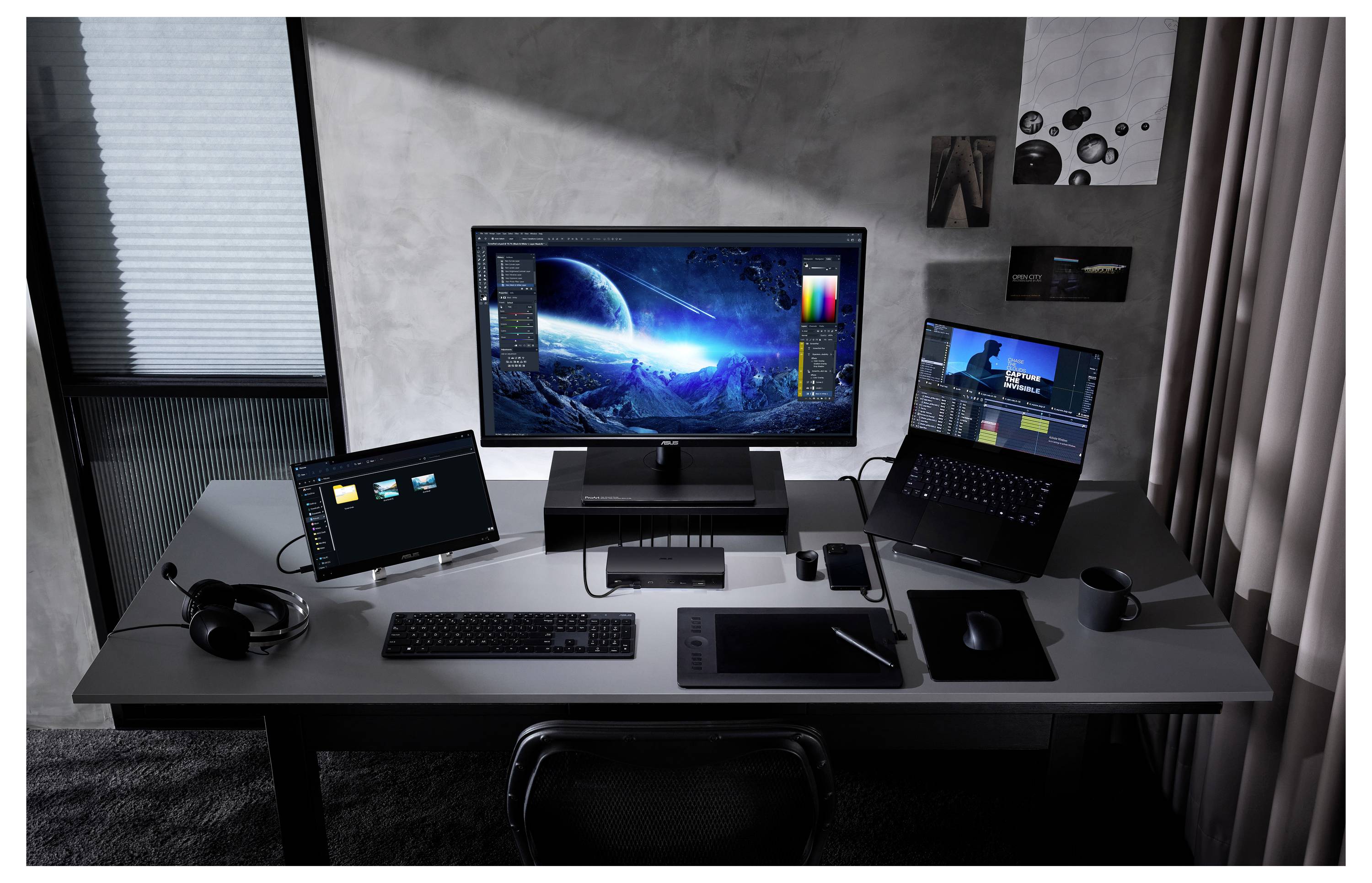 Et moderne arbejdsområde med en stor skærm, der viser et science fiction-landskab, to bærbare computere på hver sin side, et grafiktablet og et tastatur.