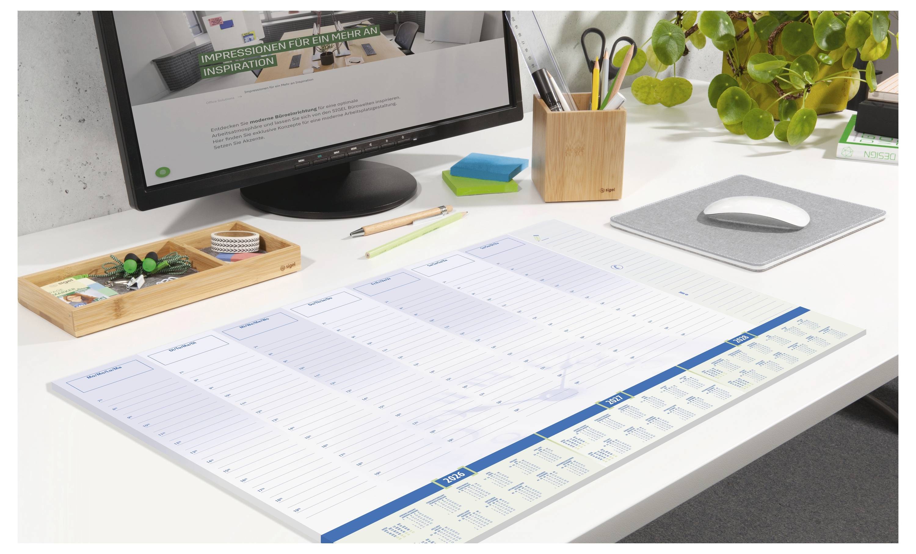 'Et ryddeligt skrivebord med en computerskærm, der viser et dokument, en ugeplanlægger, kontorartikler og en potteplante.'