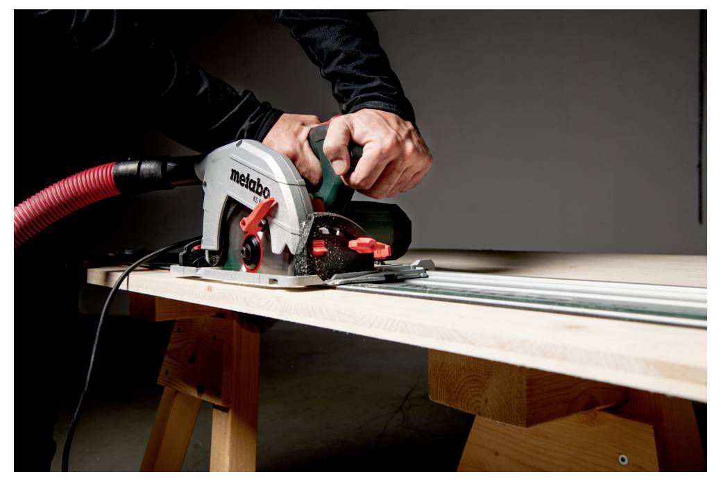 En person, der bruger en Metabo rundsav på et stykke træbræt, med fokus på præcisionsskæring. Saven er forbundet til en rød støvsugerslange til støvopsamling.