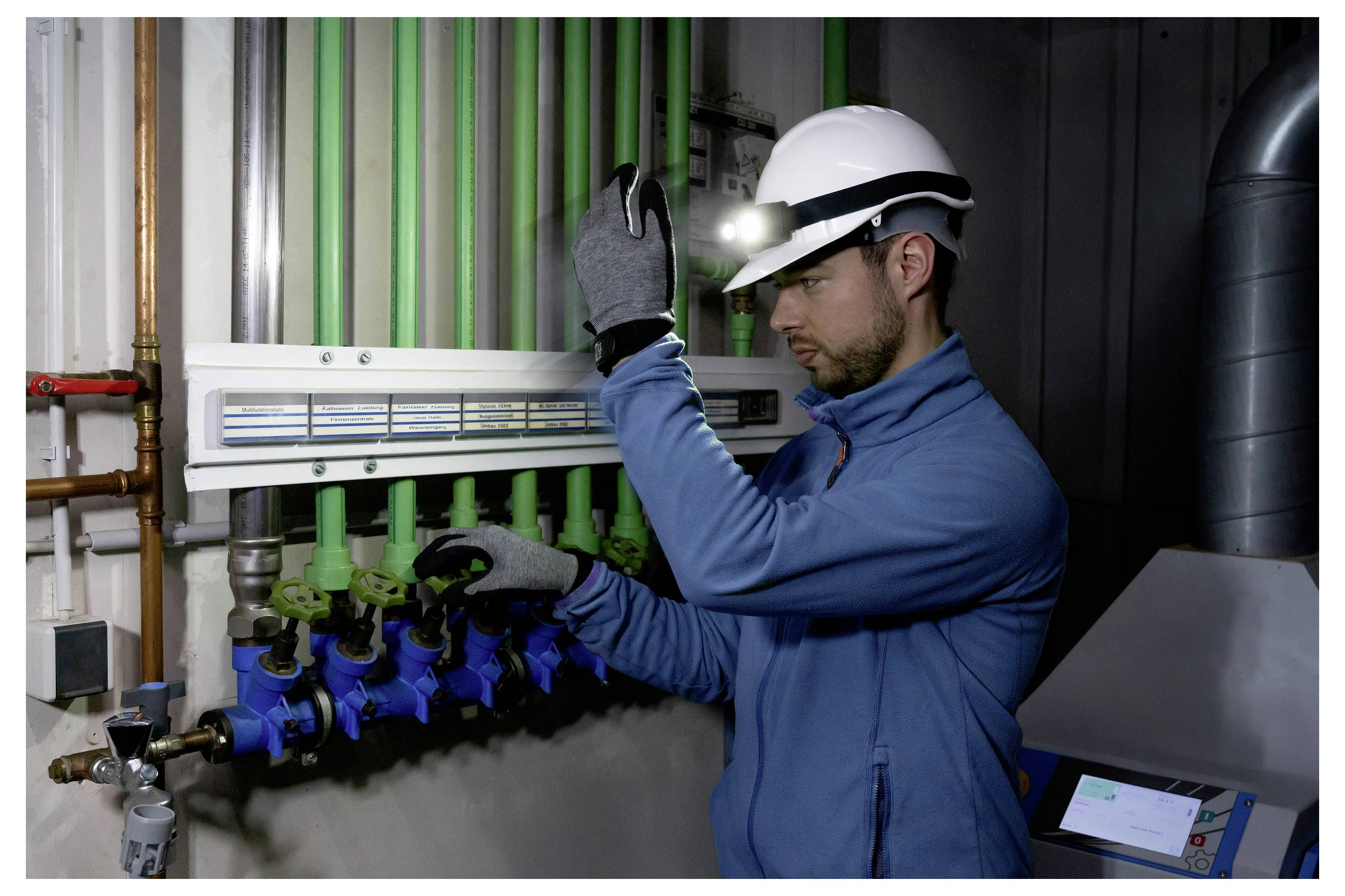 En person iført en sikkerhedshjelm og pandelampe inspicerer rør og ventiler i et industrielt miljø og justerer udstyr med en handsket hånd.