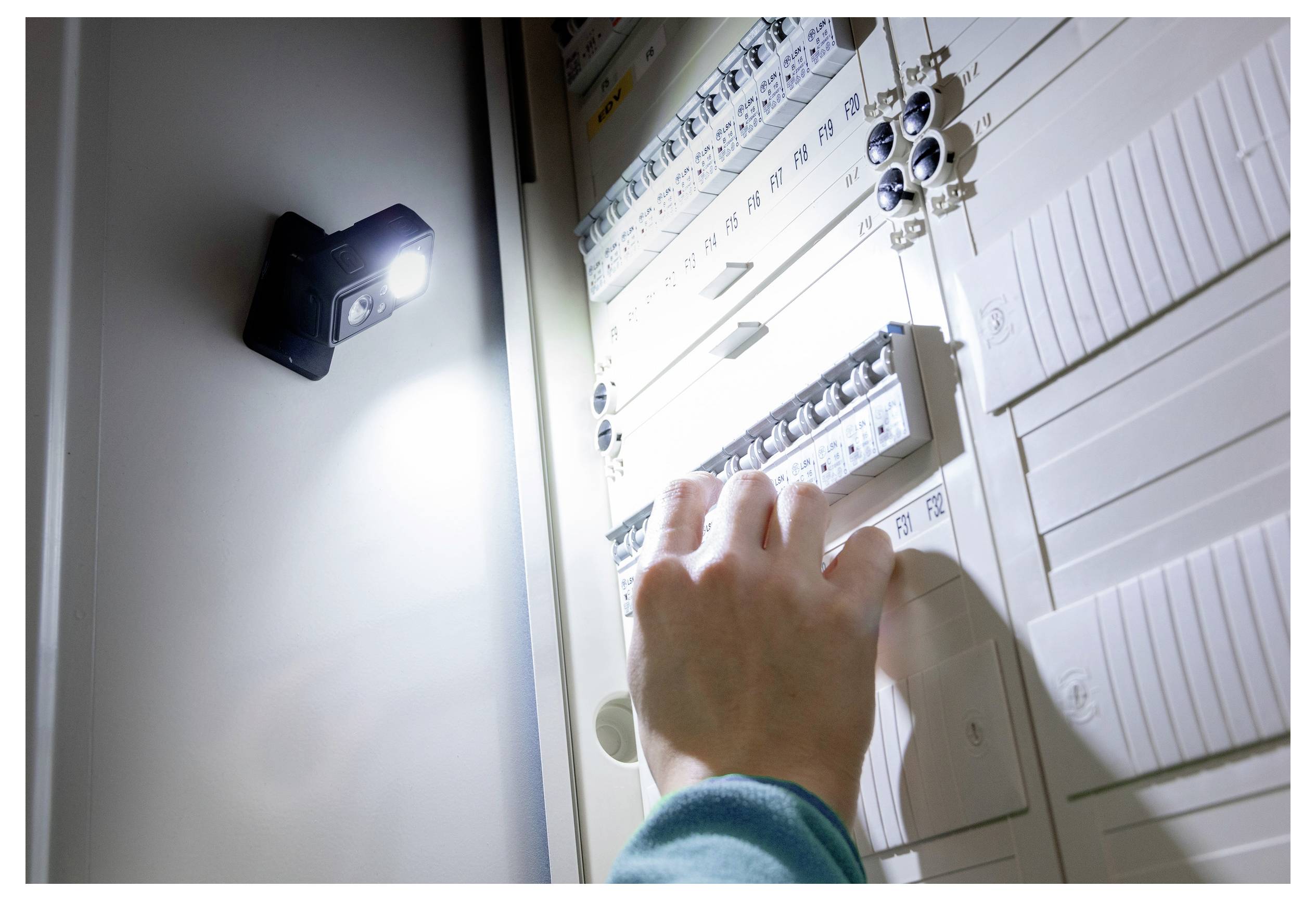 En hånd, der rækker ud for at betjene kontakter i et elektrisk panel, belyst af et lille lys, hvilket antyder vedligeholdelses- eller inspektionsarbejde.