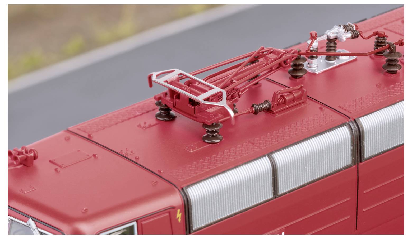 Topview af et rødt togtags model, der viser detaljerede elektriske komponenter og en pantograf mod en sløret udendørsbaggrund.