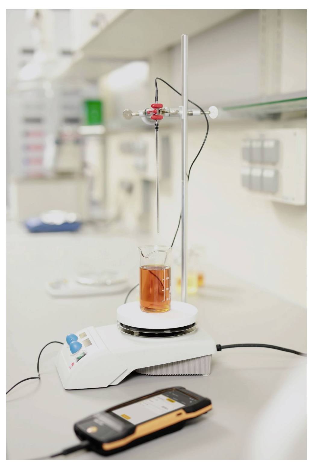 Et laboratorieopstilling med et bæger fyldt med orange væske på en magnetrører, forbundet til en sonde. En digital enhed er i forgrunden.