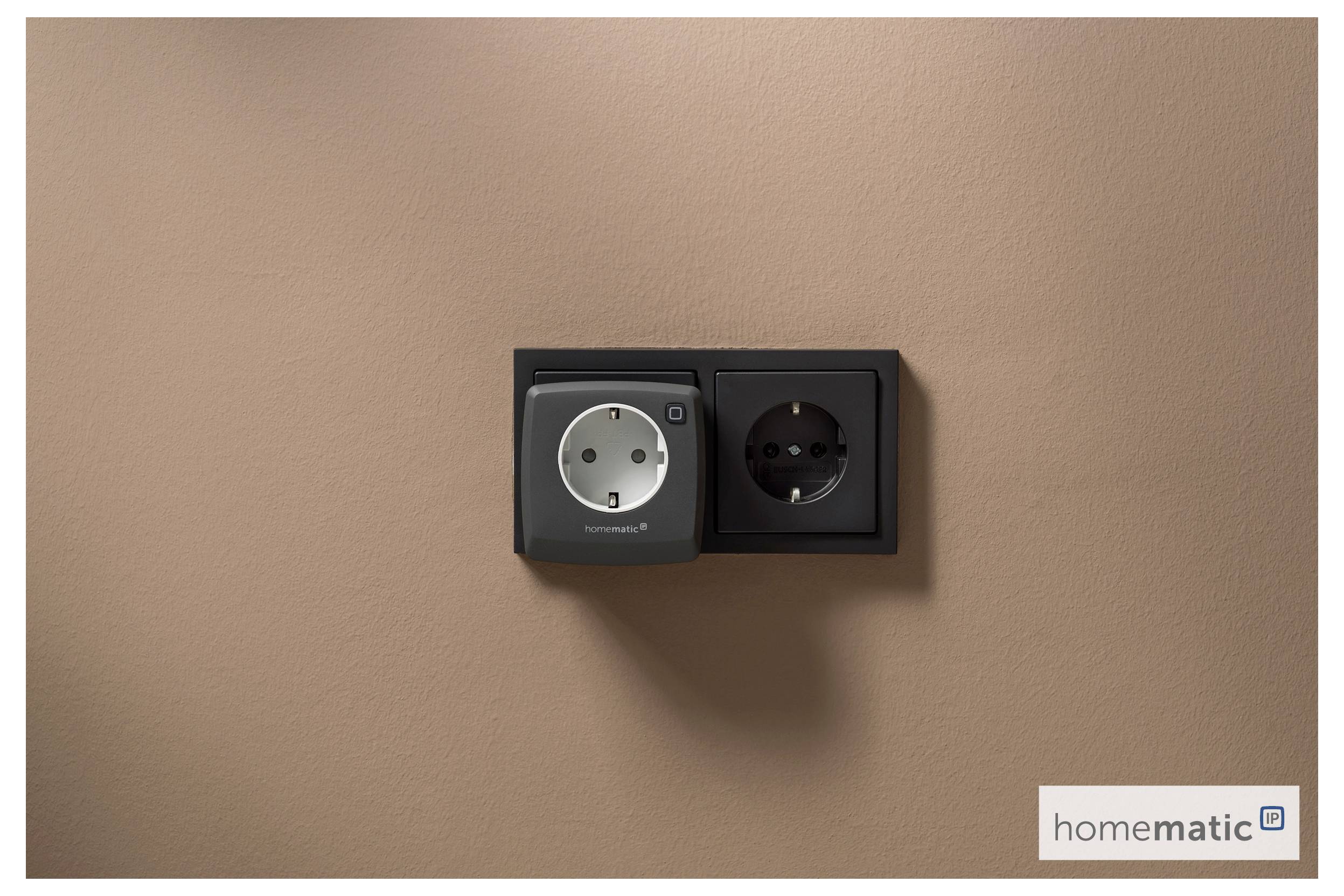 Et dobbelt stikkontakt med en elegant smart stik-adapter op ad en beige væg, mærket 'homematic' i nederste højre hjørne.