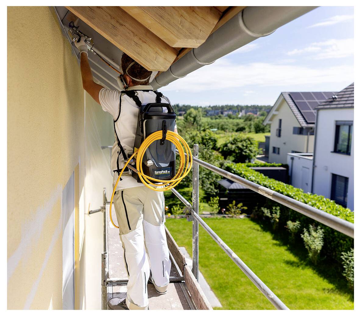 En person, der står på stilladset, maler husets facade med en malersprøjte. Huse og grønne planter er synlige i baggrunden.