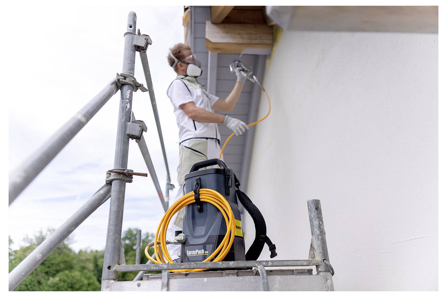 'En person iført maske og handsker bruger en malingsprøjte på en husfacade, mens vedkommende står på stilladset. En slange og udstyr er placeret i nærheden.'
