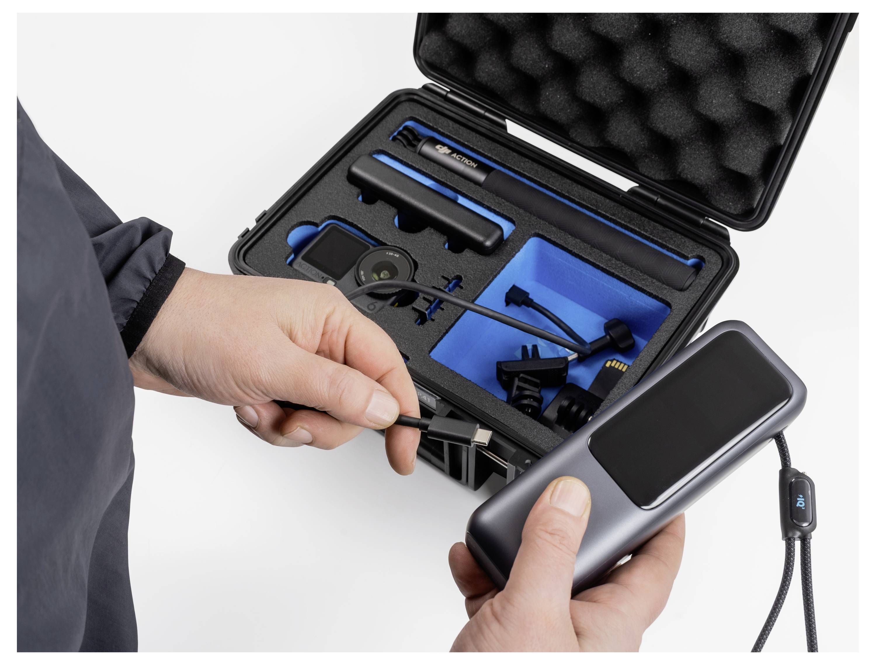 En person holder en bærbar elektronisk enhed med en skærm og et kabel ved siden af et åbent sort etui med organiserede rum.