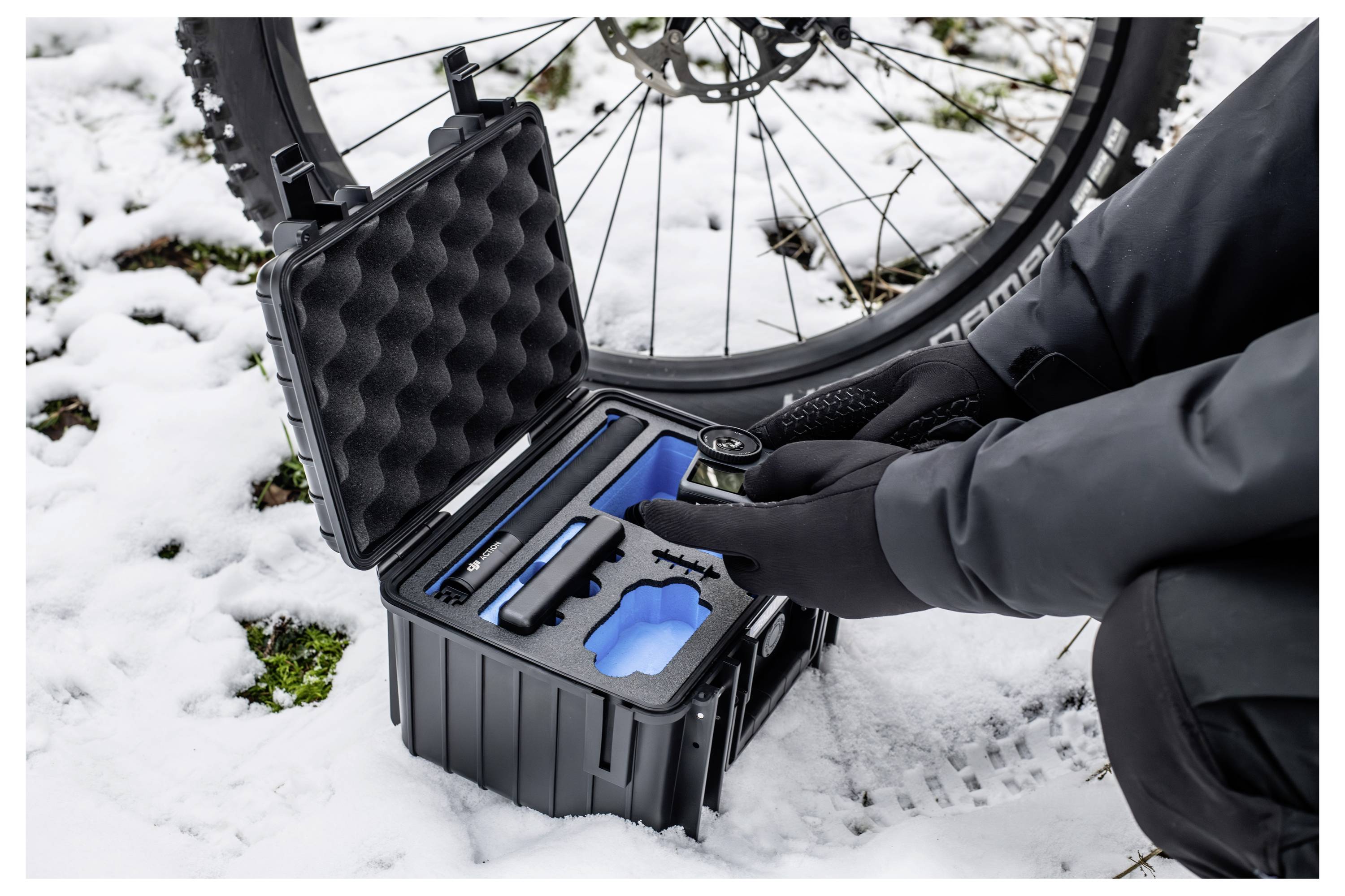 En person i vinterklæder knæler ved siden af en cykel og holder en komponent fra et åbent værktøjskasse. Jorden er dækket med sne.