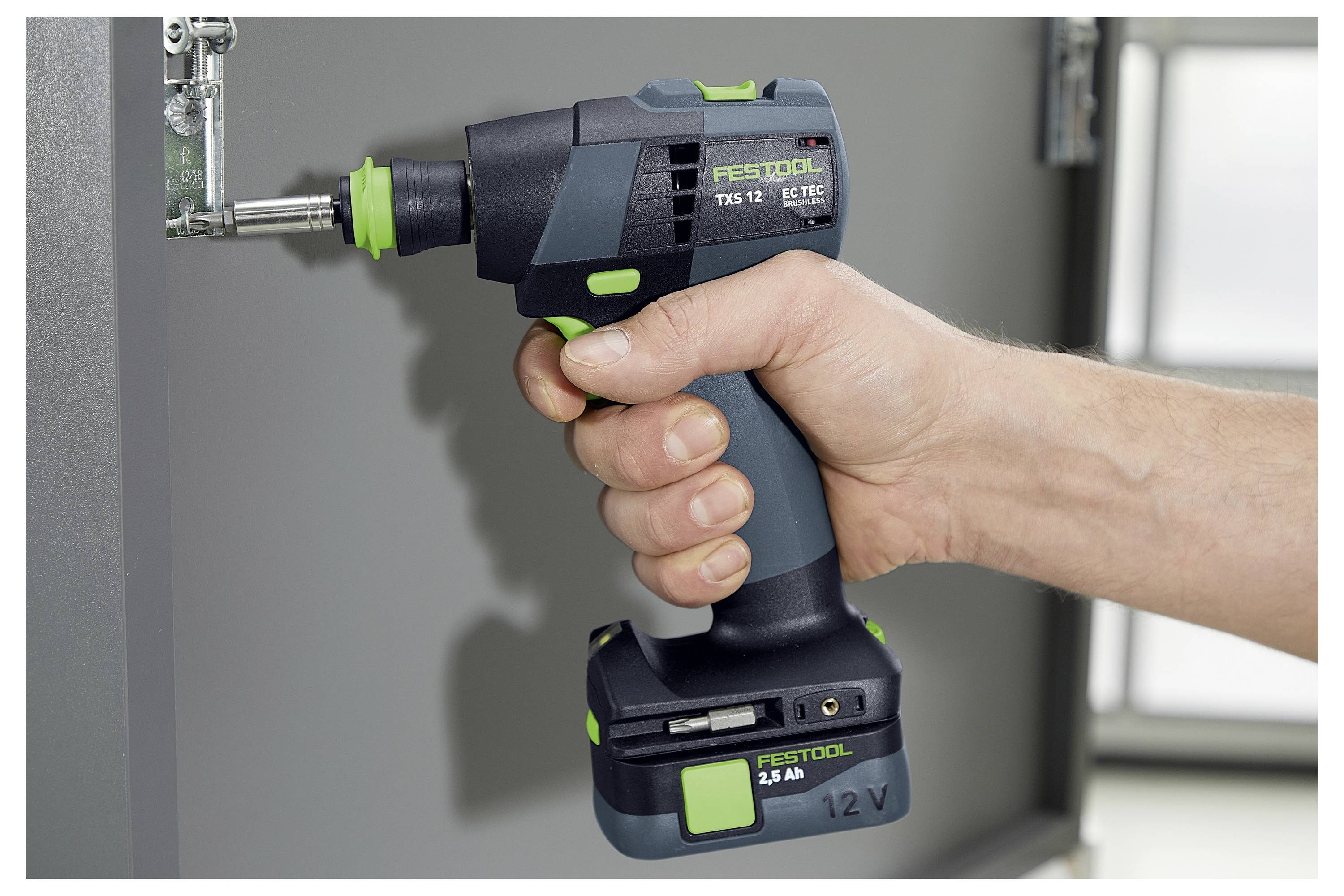 'En person bruger en FESTOOL 12V elektrisk skruetrækker til at stramme en skrue på et hængsel fastgjort til et møbel.'