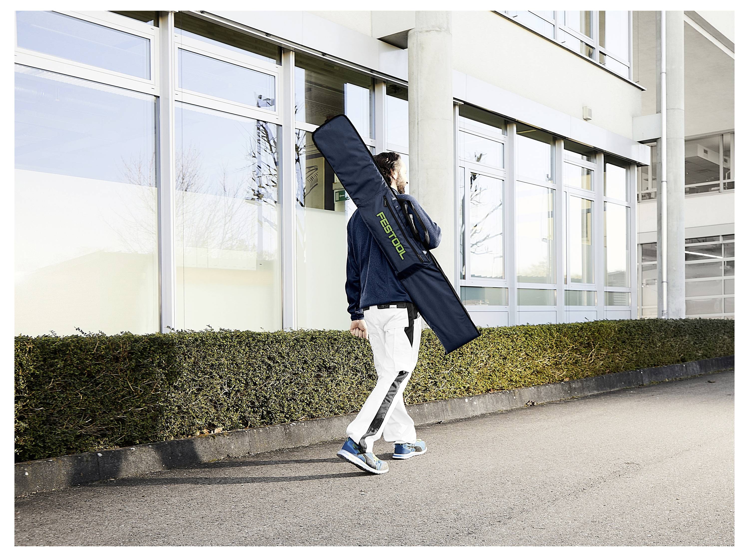 En person går langs en fortov og bærer en lang, sort taske over skulderen. Tasken har 'FESTOOL' skrevet med grønne bogstaver.