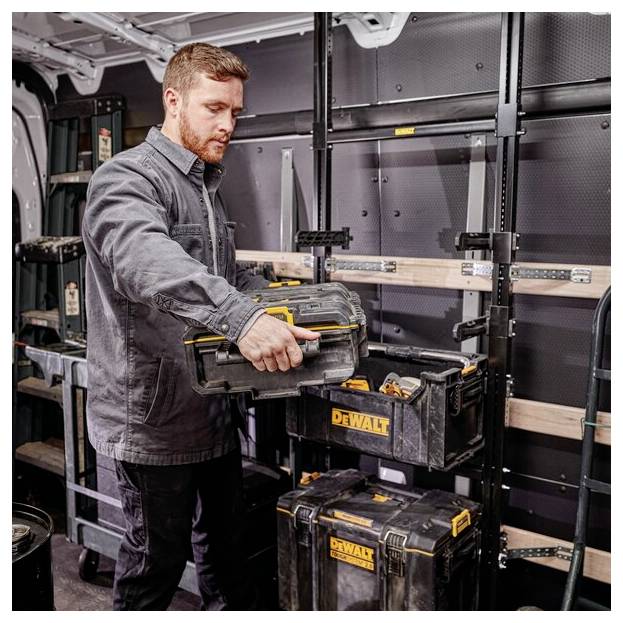 En person i en arbejdsbil organiserer værktøjskasser mærket 'Dewalt' på metalreolfer, iført en grå arbejdsjakke.