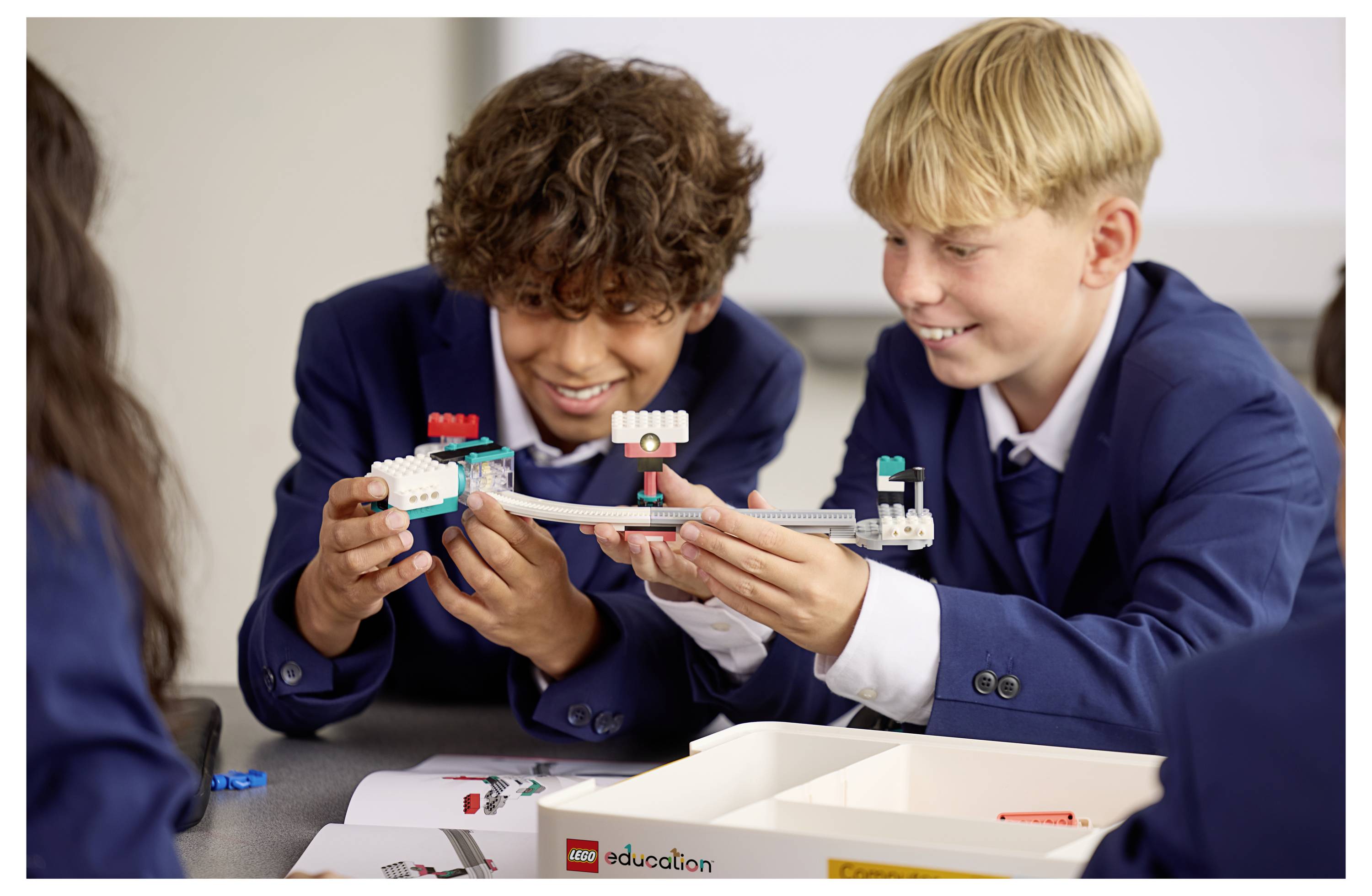 To studerende i skoleuniforms, der bygger en robot ved hjælp af Lego Education-sæt, smiler og samarbejder ved et klasseværelsesbord.