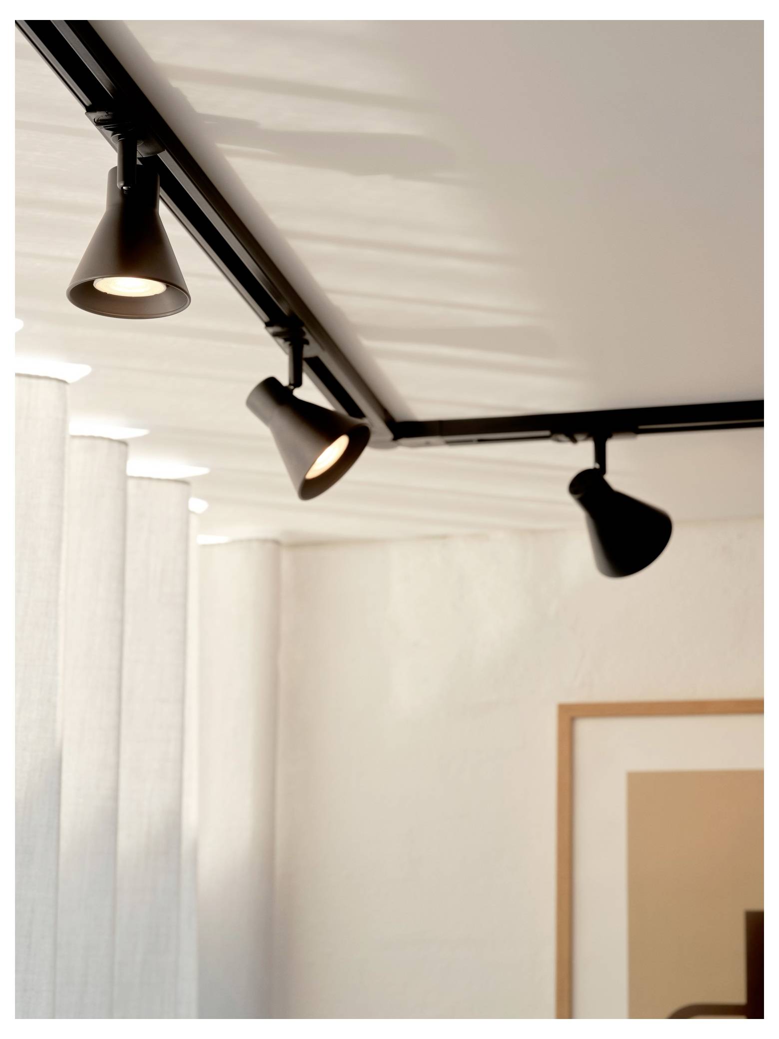 Sporrelampe med tre justerbare sorte spotlights på et hvidt loft, der belyser et minimalistisk rum med lette grå gardiner.
