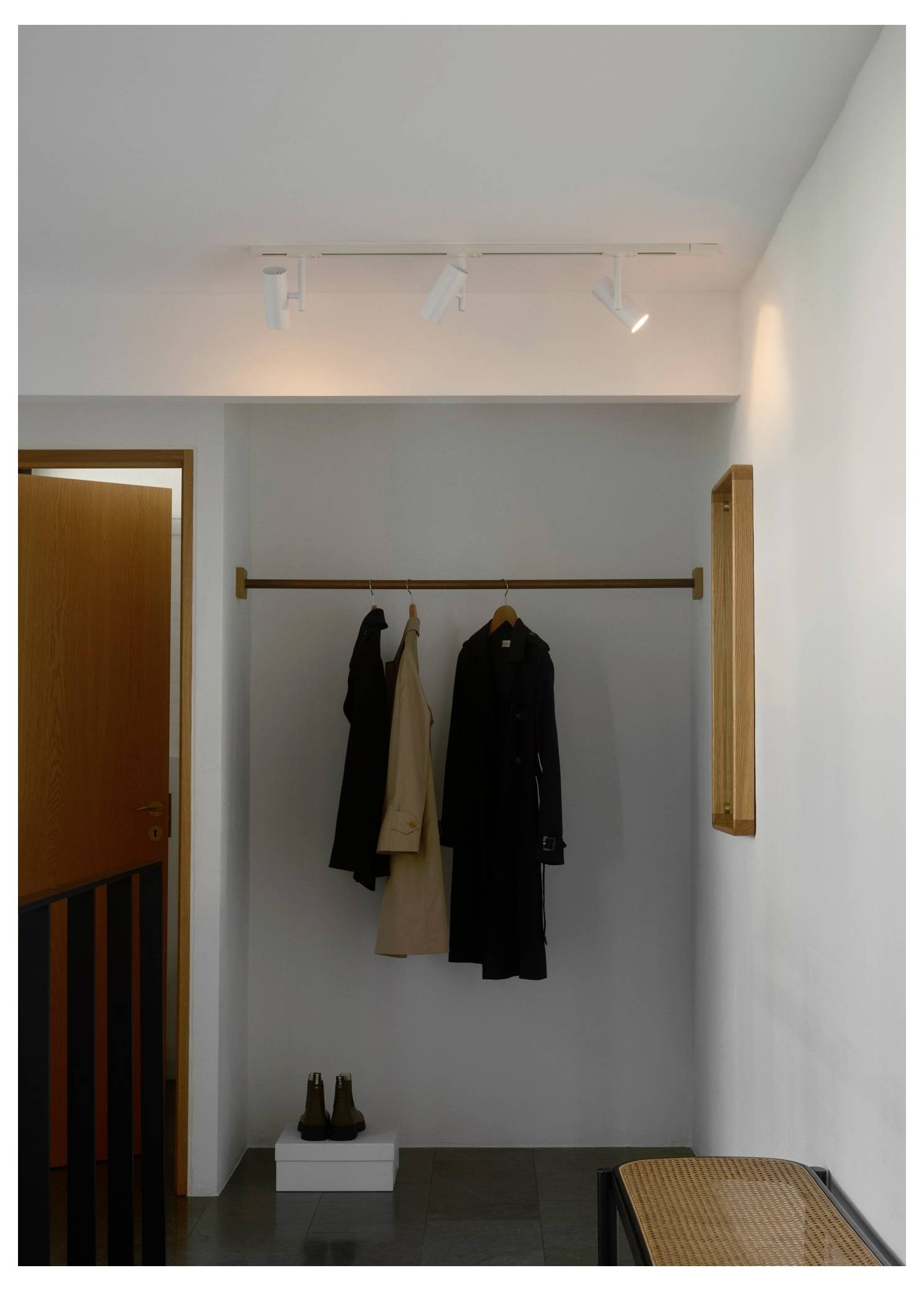 En minimalistisk entré med en træ-kappestativ, der holder to frakker. Et par sko er pænt placeret på en lille hvid platform nedenunder.