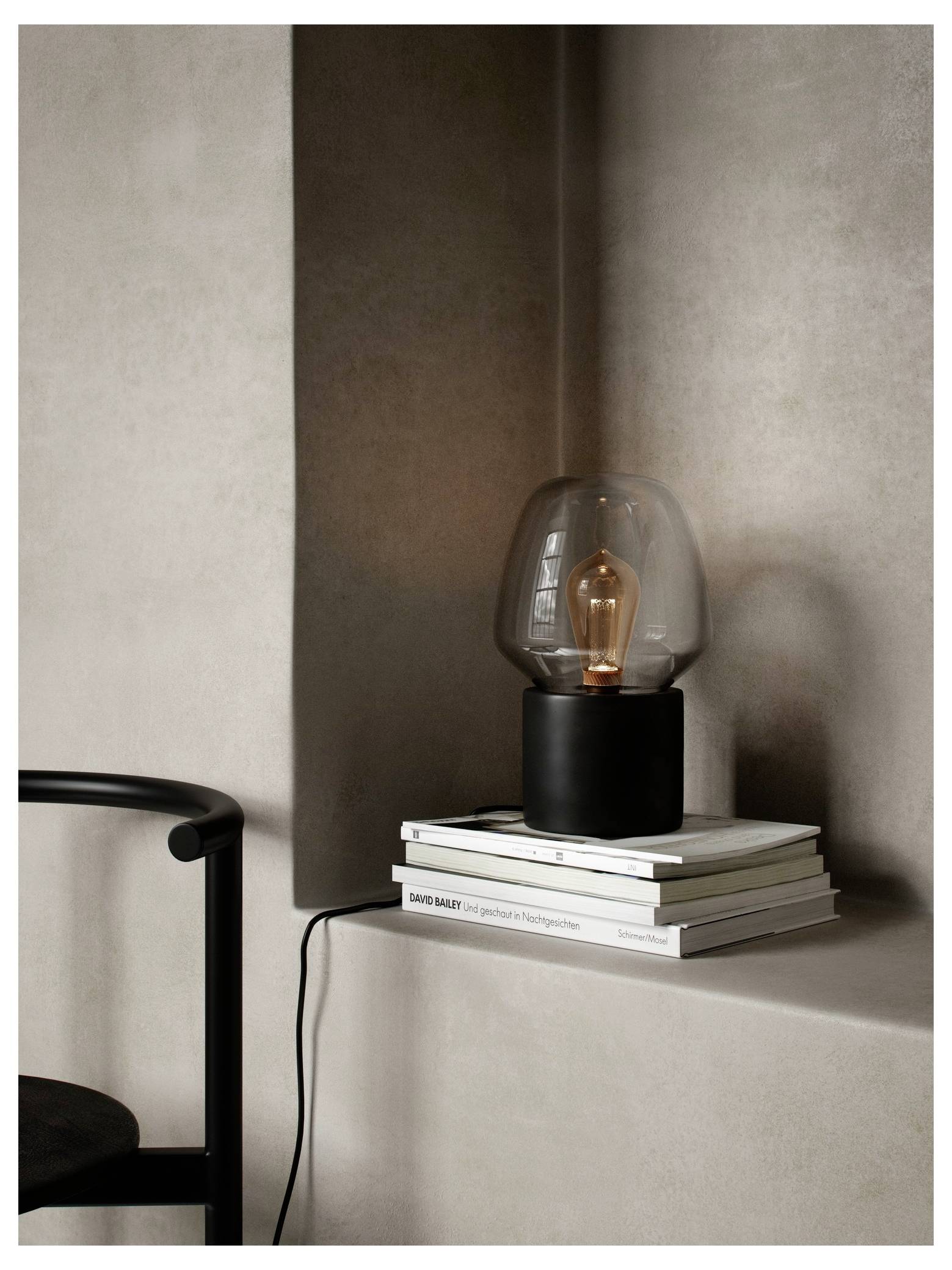 En moderne glasplampe står på staklet bøger i et minimalistisk rum med neutrale toner, ved siden af en sort stol.