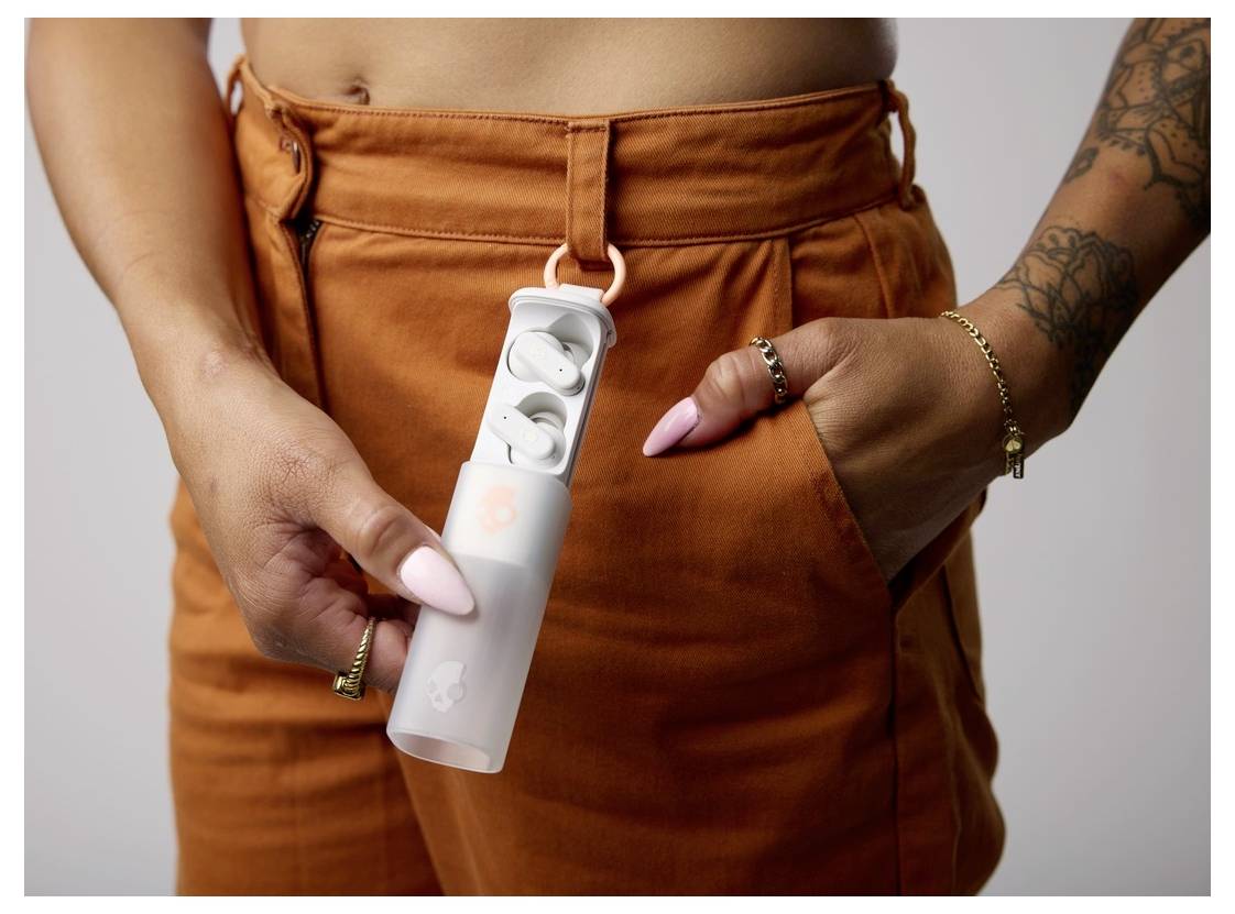 En person, der holder en lille etui med trådløse øretelefoner i det, iført orange bukser og ringe på fingrene.