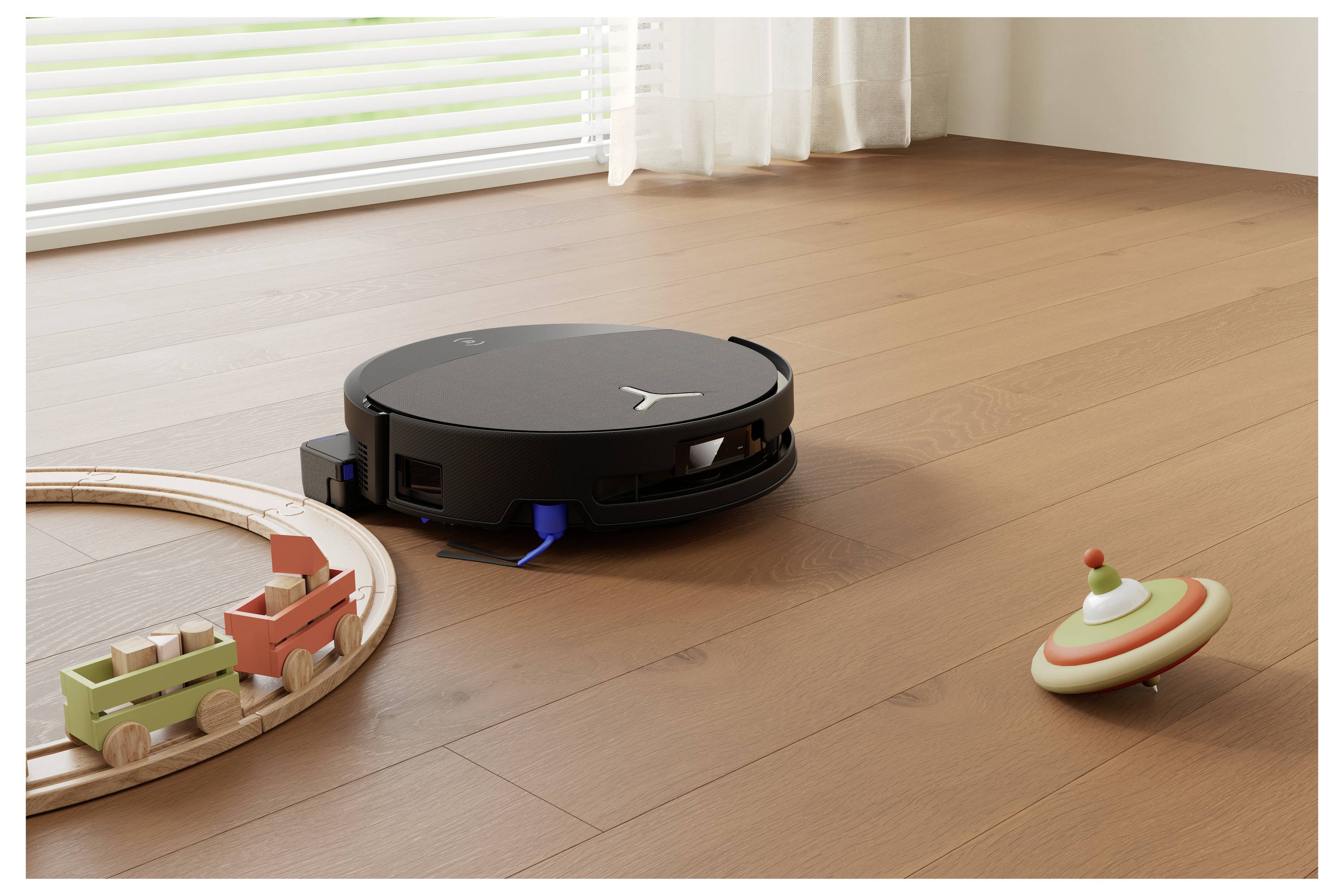 En sort cirkulær robotstøvsuger, der renser et trægulv nær et legetøjstog på skinner og en snurrende top i et solrigt rum.