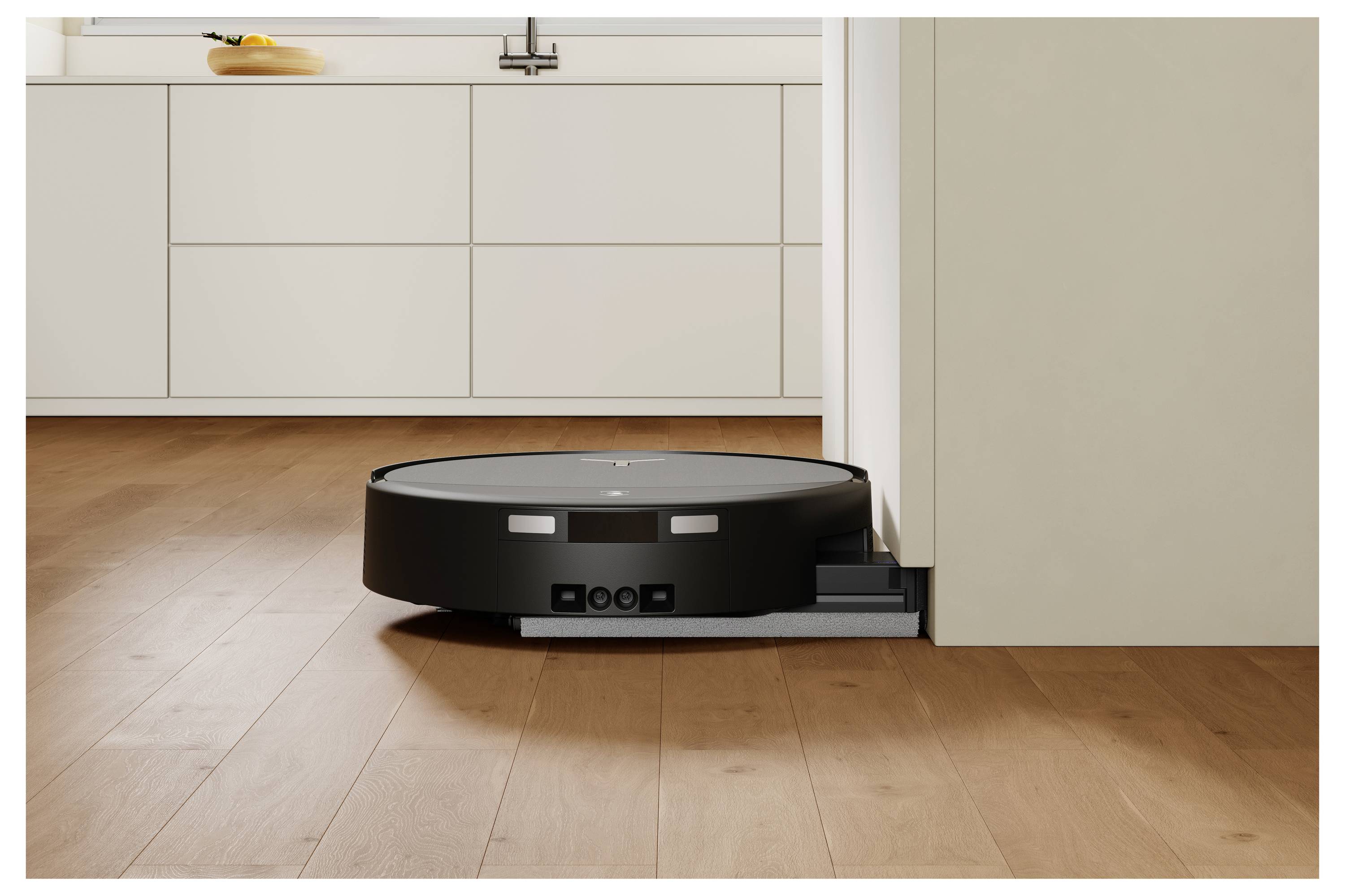 En sort robotstøvsuger er ved at dokke sig selv mod en hvid fodpanel i et rum med træfloor.