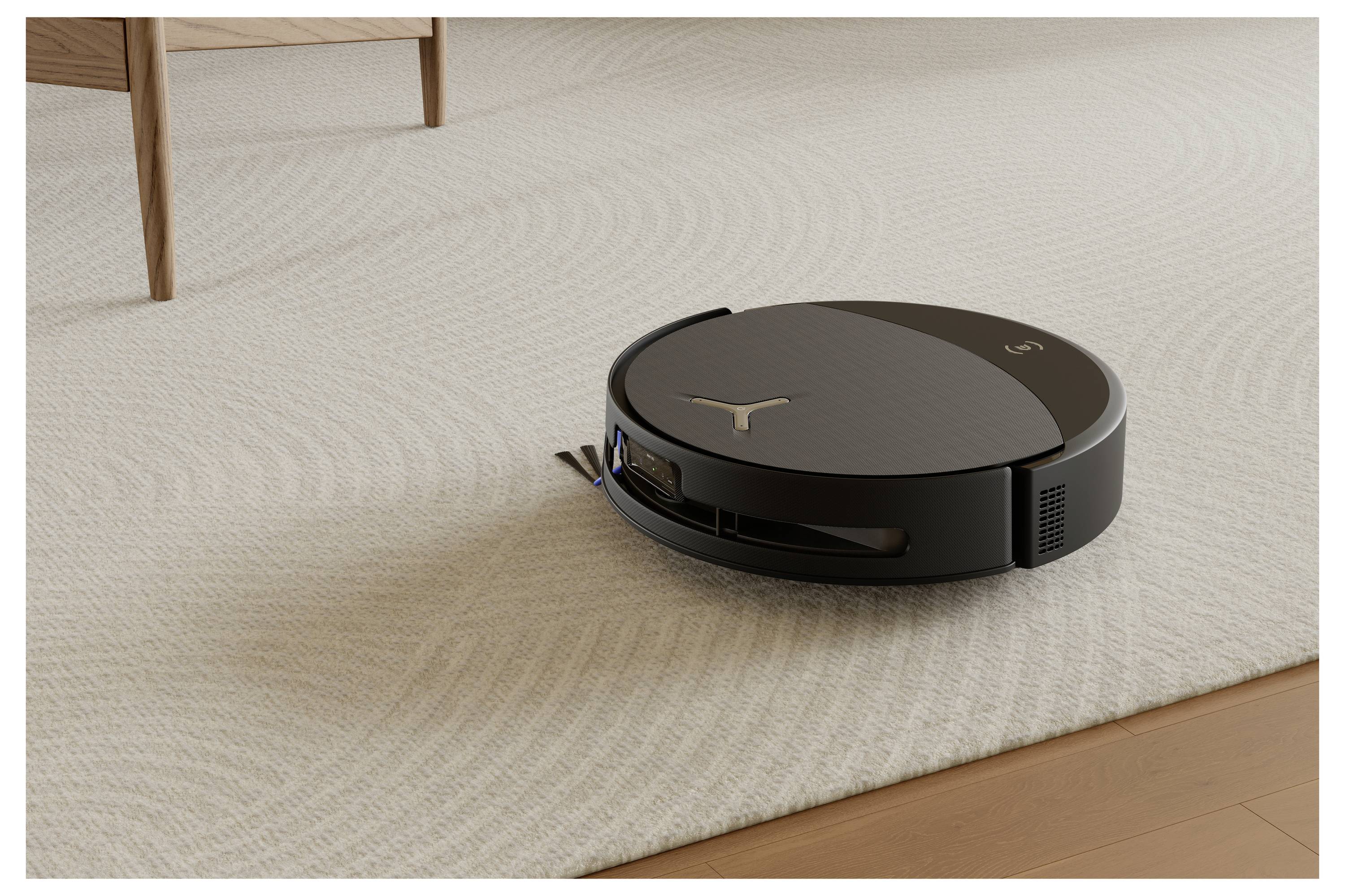 En sort robotisk støvsuger på et tæppe, ved siden af et møbel af træ. Den rengør autonomt i et hjemmemiljø.