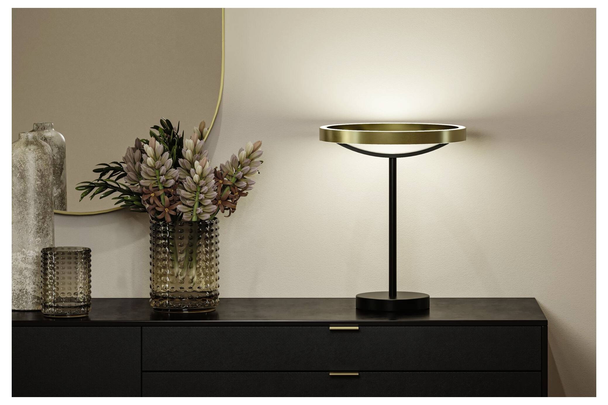 Moderne sort bordlampe med en lysende ringformet skærm på en mørk kommode ved siden af en glaskrukke med tørrede blomster og en stearinlysholder.
