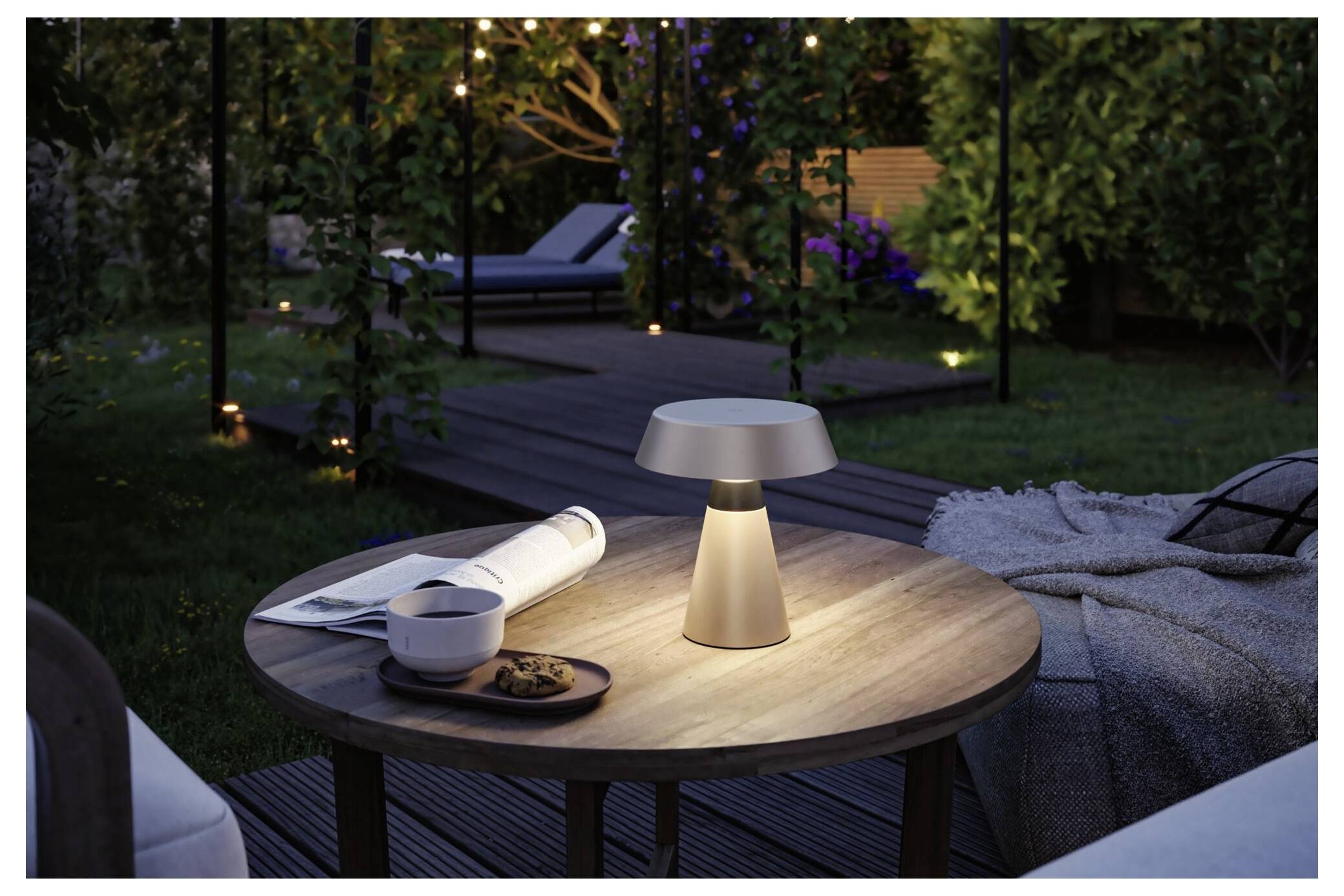 Bærbar bordlampe, der oplyser et udendørs patibord ved siden af et rullet magasin, en skål og et tæppe i en have i skumringen.