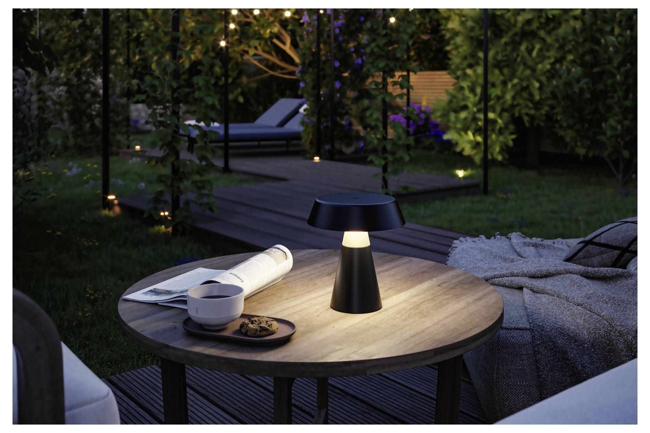 Et oplyst udendørs patioborg med en moderne lampe, kop, magasin og småkager ved siden af en polstret loungestol i skumringen.