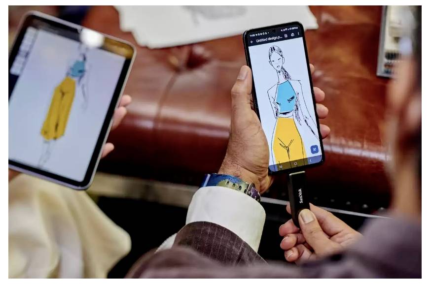 To personer sammenligner modedesign på en tablet og en smartphone, der viser en farverig tøjskitse på telefonens skærm.