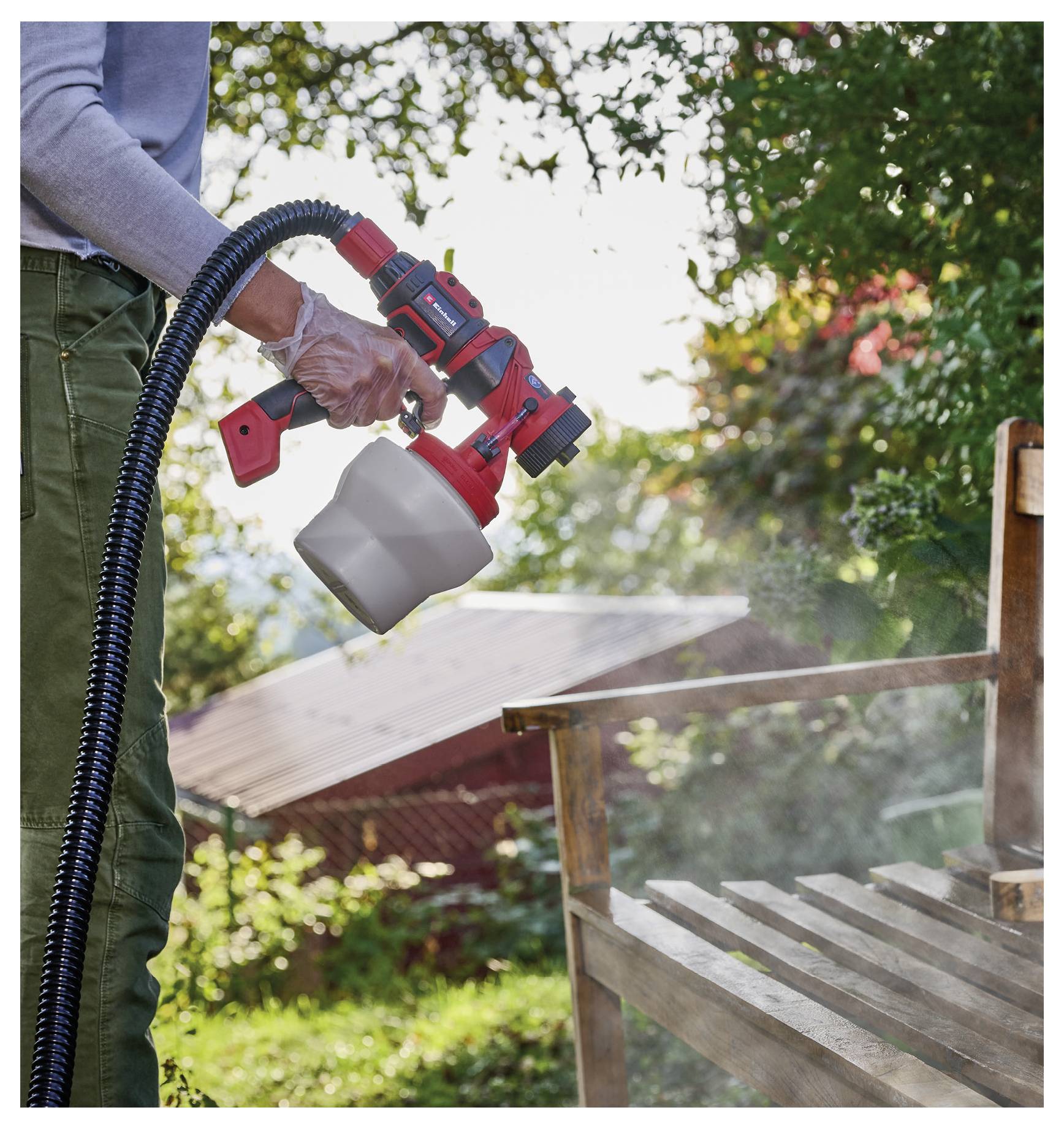 En person, der bruger en håndholdt malesprøjte til at påføre farve på en træ-udendørsbænk eller stol i en baghave, med synligt spray-tåge.