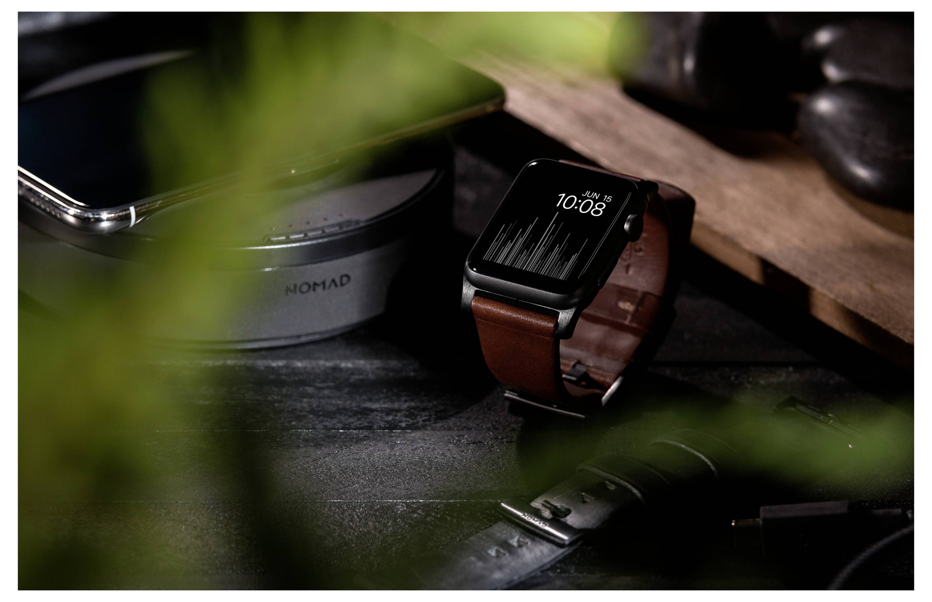 Apple Watch med brunt læderrem, der viser 10:08 på et mørkt skrivebord ved siden af en telefon, delvis skjult af uskarpe blade.