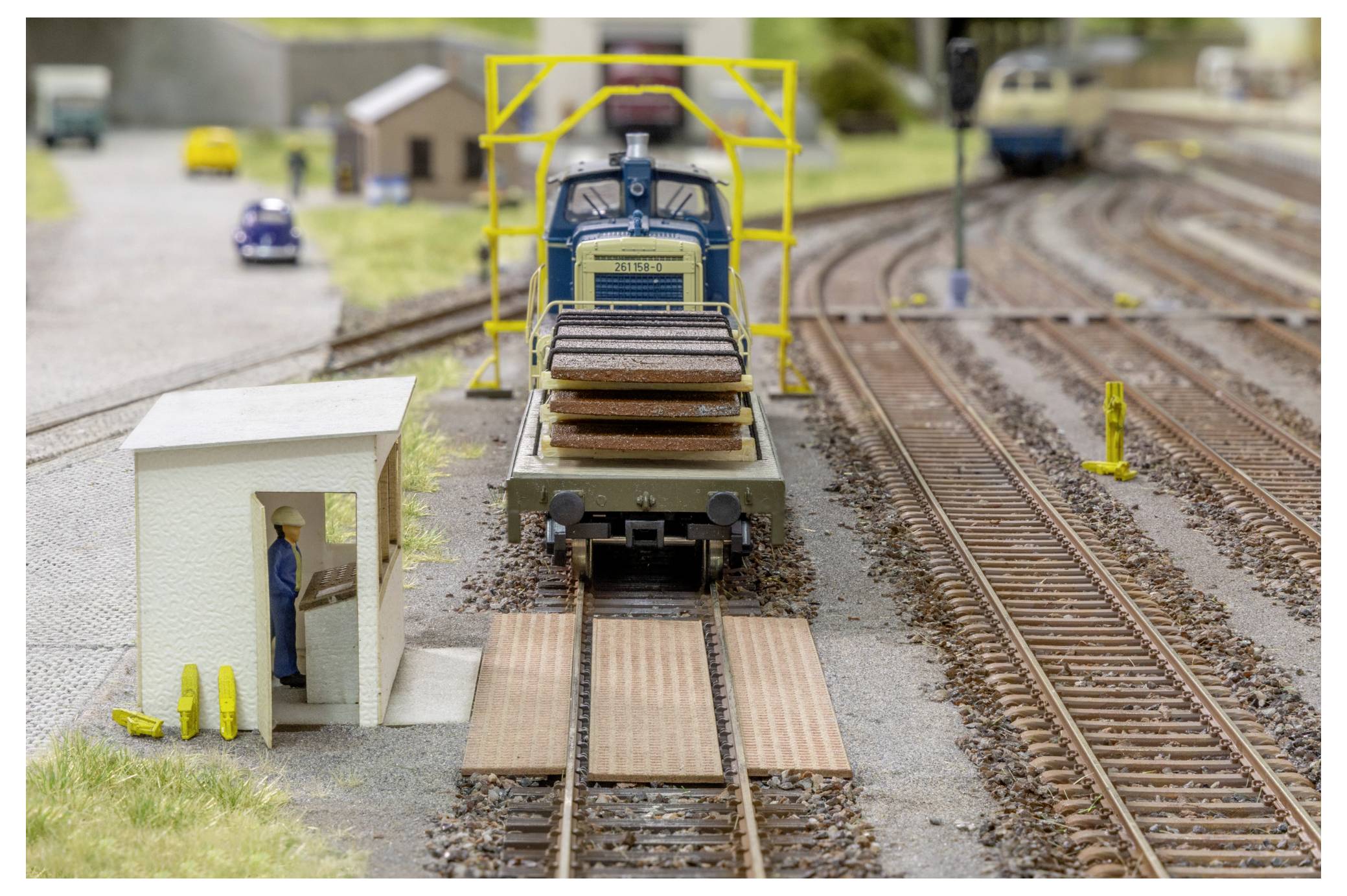 Modeltoget på skinner nærmende sig et lille jernbaneovergang, med en miniatyrarbejder ved et sporskur og flere jernbanespor i baggrunden.