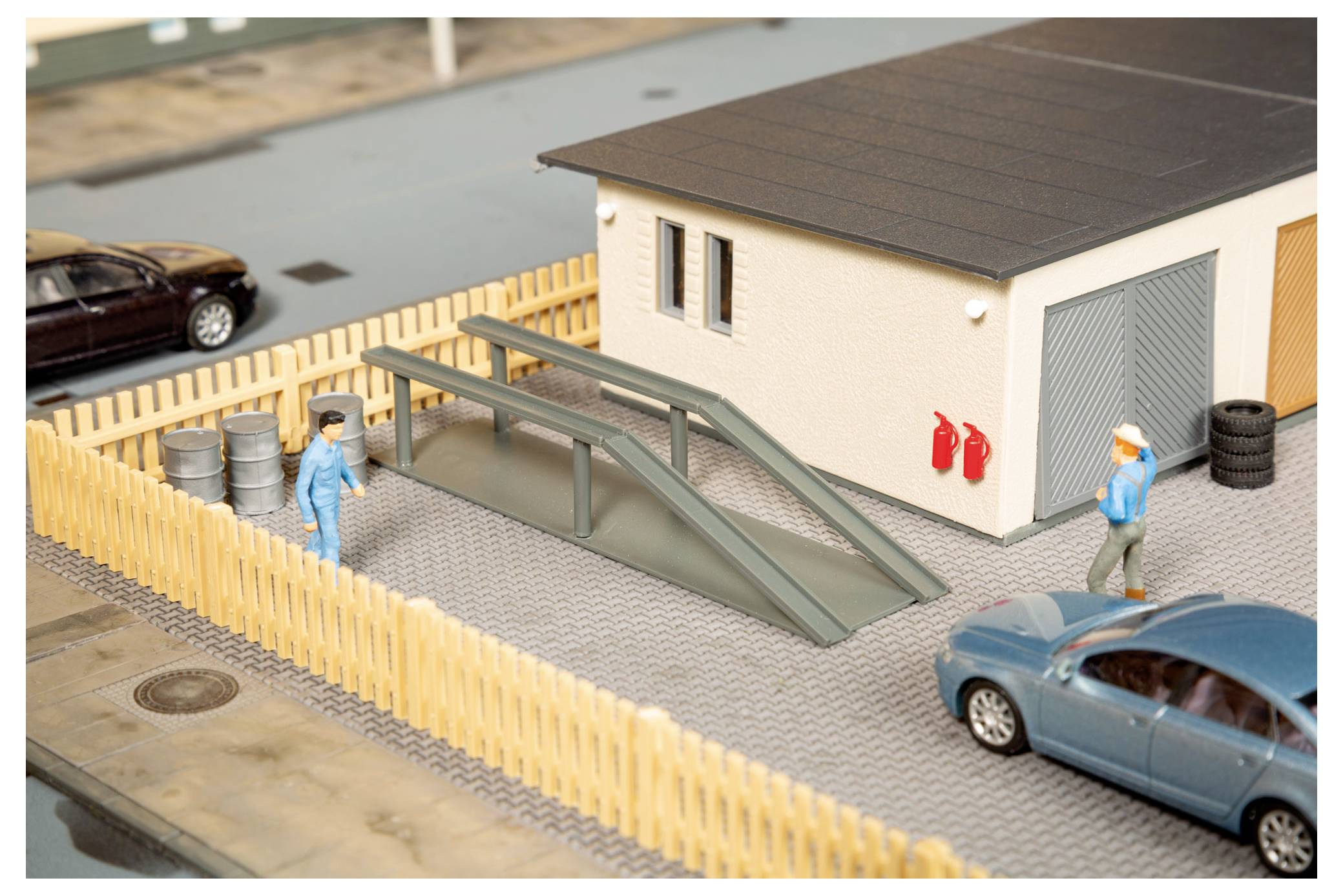 Miniatyrscene ved vejsiden med en cremefarvede bygning, indhegnet grund, rampestruktur, to arbejderfigurer, parkerede biler og røde brandslukningsapparater.