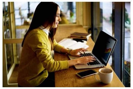 Kvinde, der arbejder på en bærbar computer ved et cafébord ved et vindue, med en telefon og en kaffekop ved siden af hende.