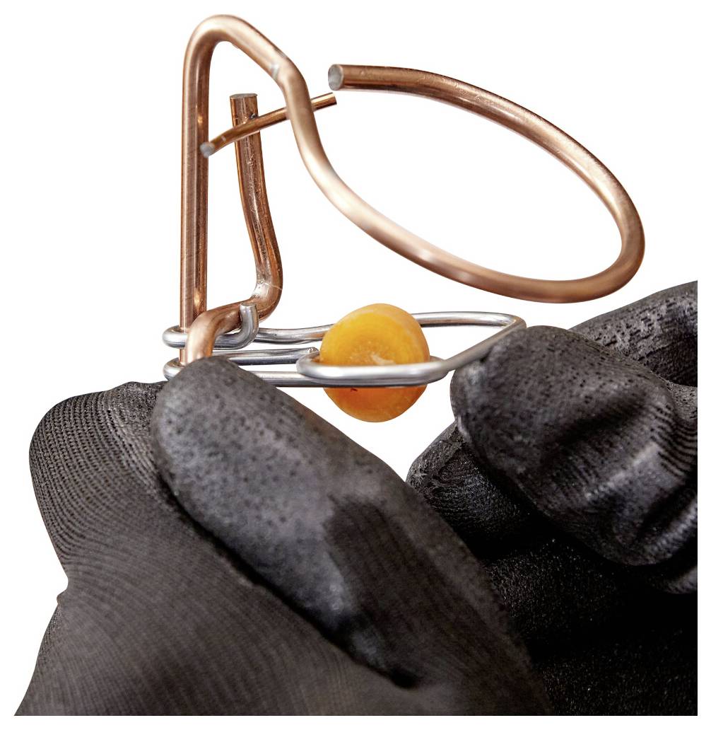 Handsker på hænder, der holder en fælde af kobber- og sølvtråd med en orange perle, hvilket antyder et close-up af smykkefremstilling eller trådarbejde.