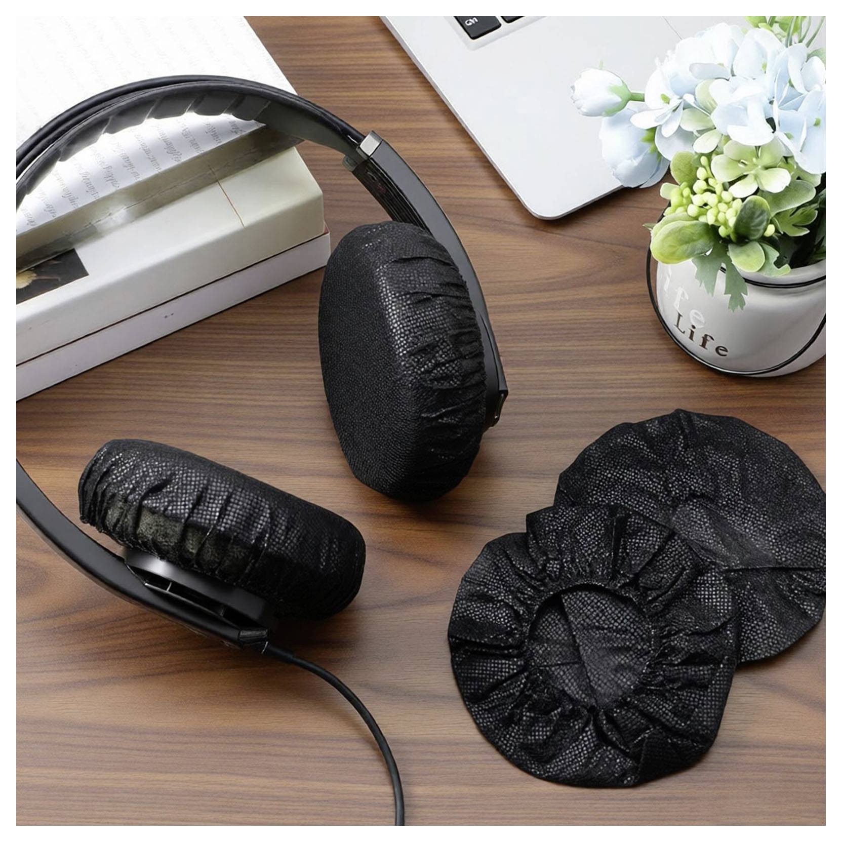 Sorte over-ear høretelefoner med udskiftelige ørepolstringer på et træbord ved siden af en bog, bærbar computer og en blomsterkrukke.